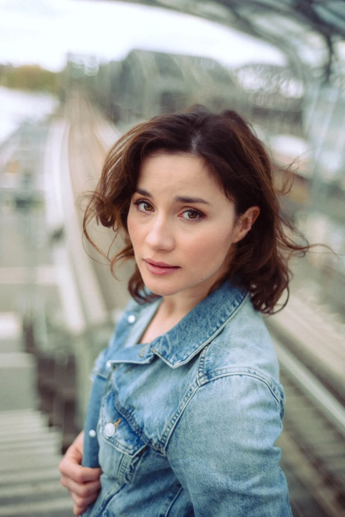 Jeannine Michèle Wacker mit braunen, gewellten Haaren trägt eine Jeansjacke und schaut in die Kamera, im Hintergrund eine weiße Brücke oder eine moderne Glasstruktur.