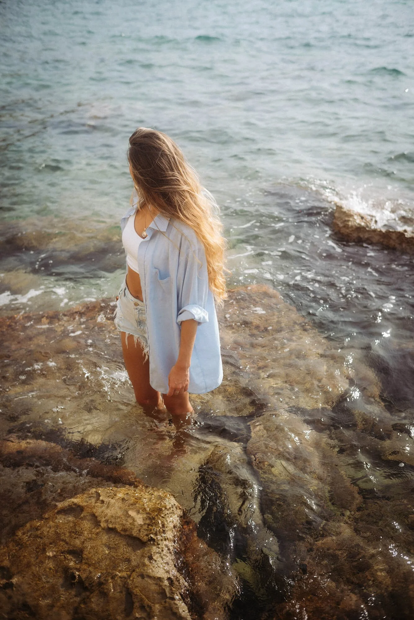 Frau mit langen Haaren in einer Bluse und Shorts steht im Wasser am Strand, blickt aufs Meer.