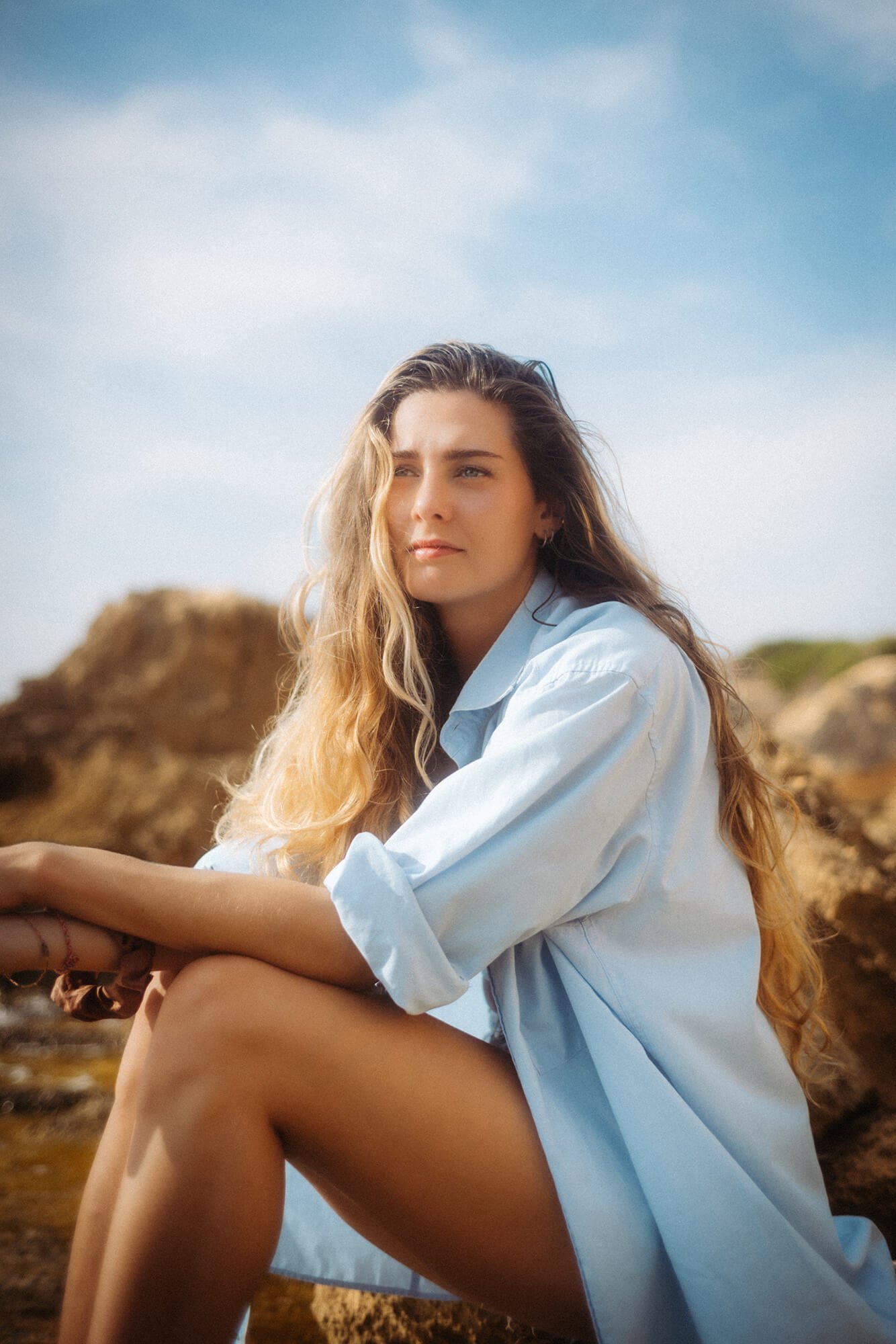 Junge Frau sitzt am Strand von Mallorca, trägt eine offene, helle Bluse und blickt nachdenklich in die Ferne, mit Felsen und Himmel im Hintergrund.