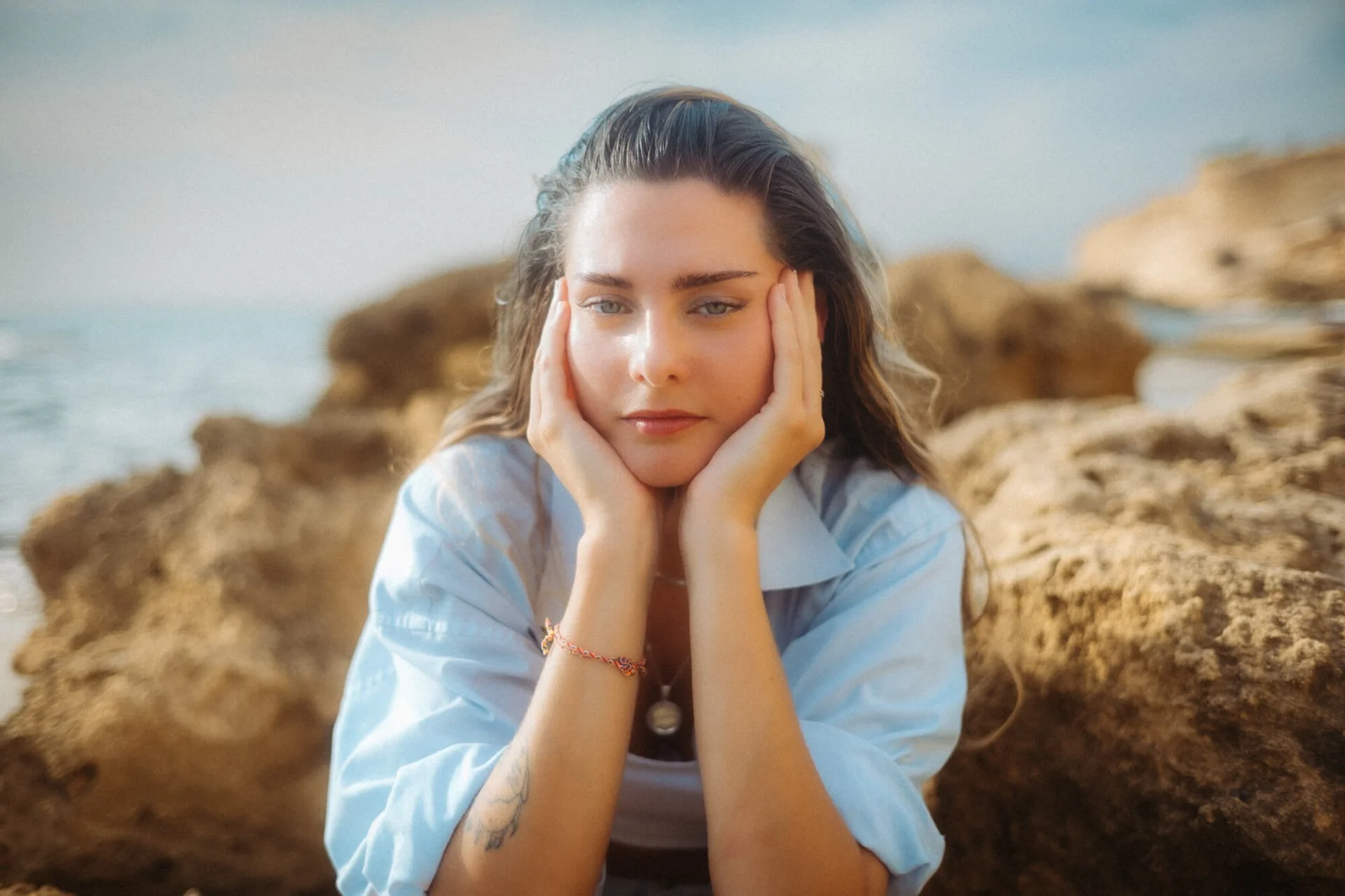 Junge Frau sitzt nachdenklich an einem Strand mit großen Steinen, Blick nach unten, Haare im Wind, blauer Himmel im Hintergrund.