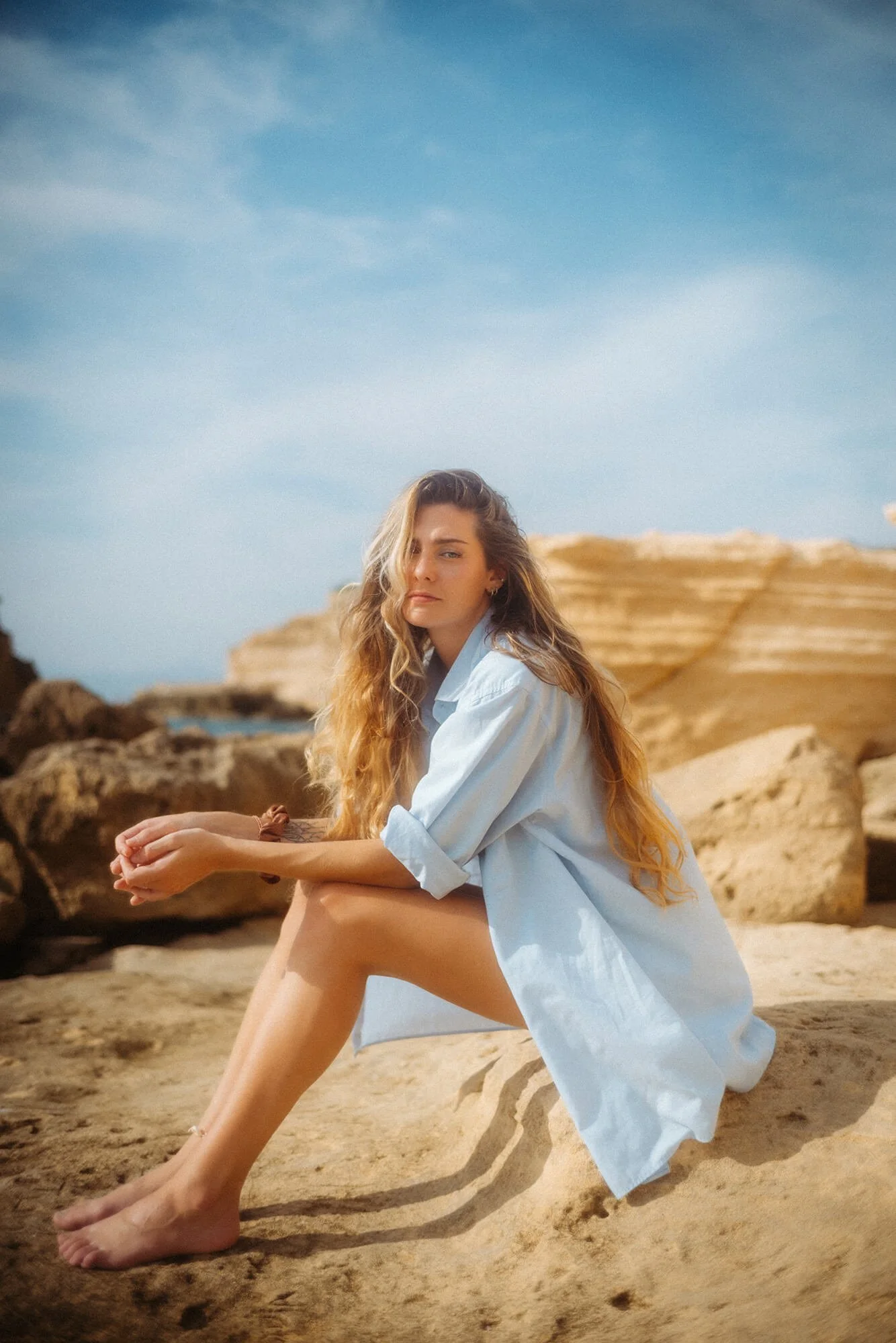 Junge Frau sitzt barfuß auf einem Sandfelsen am Strand, trägt ein lockeres weißes Hemd, mit langen, gewellten, hellbraunen Haaren, blaue Himmel mit wenigen Wolken im Hintergrund.