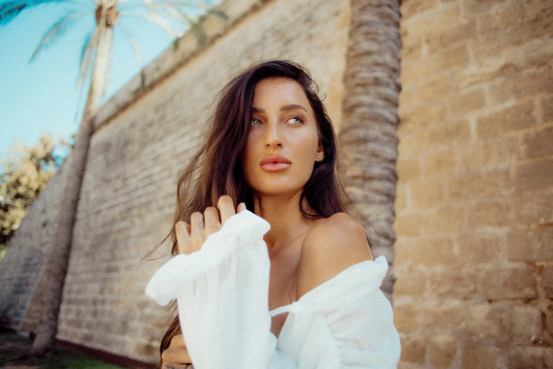 Eine junge Frau mit langen braunen Haaren steht vor einer Steinmauer und einer Palme, die bei Sonnenlicht fotografiert wurde.