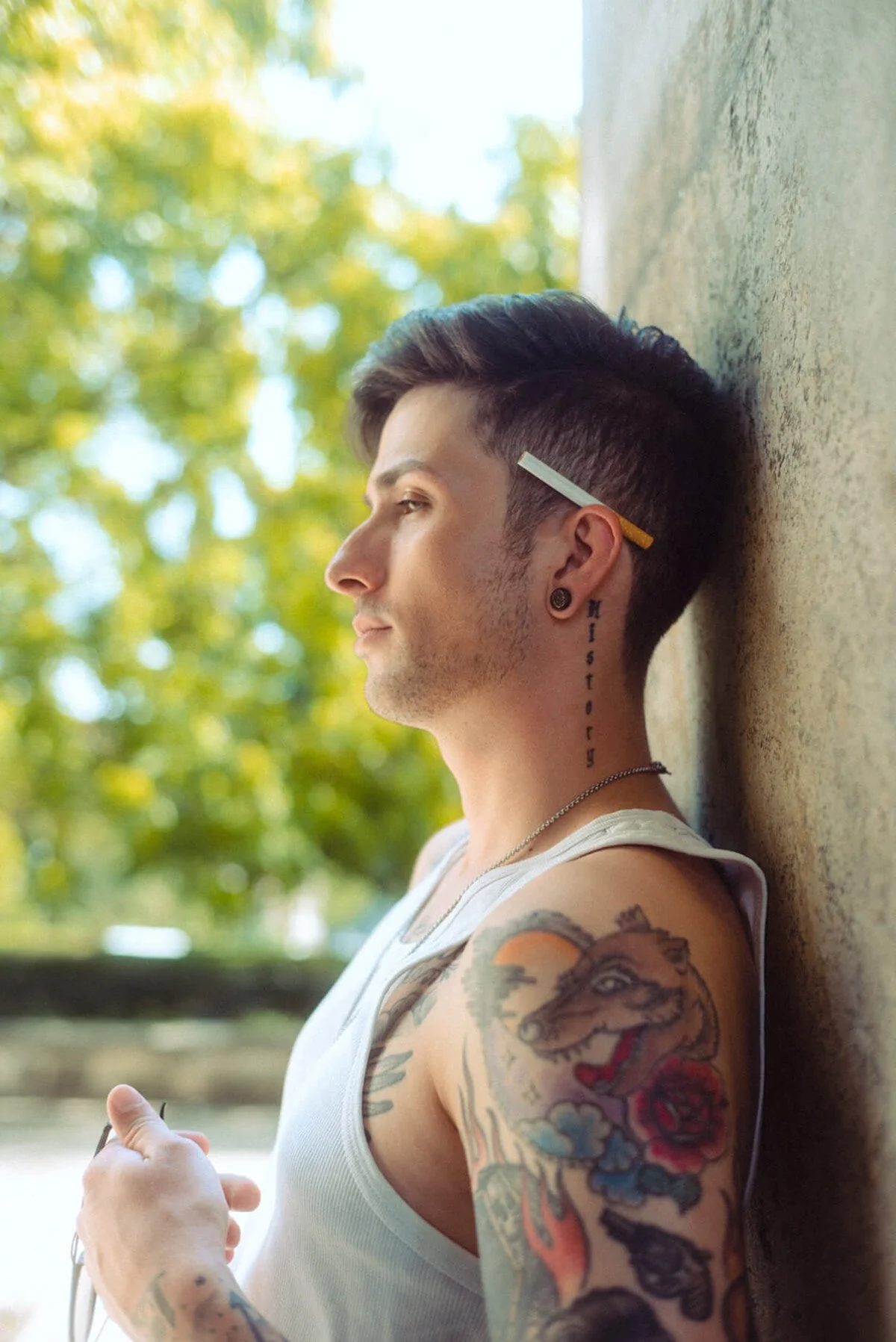 Benjamin Mittelstädt von Berlin Tag und Nacht mit Tattoos, Piercing und Zigarettenspitze, lehnt an einer Wand im Freien, während er die Sonnenbrille hält.