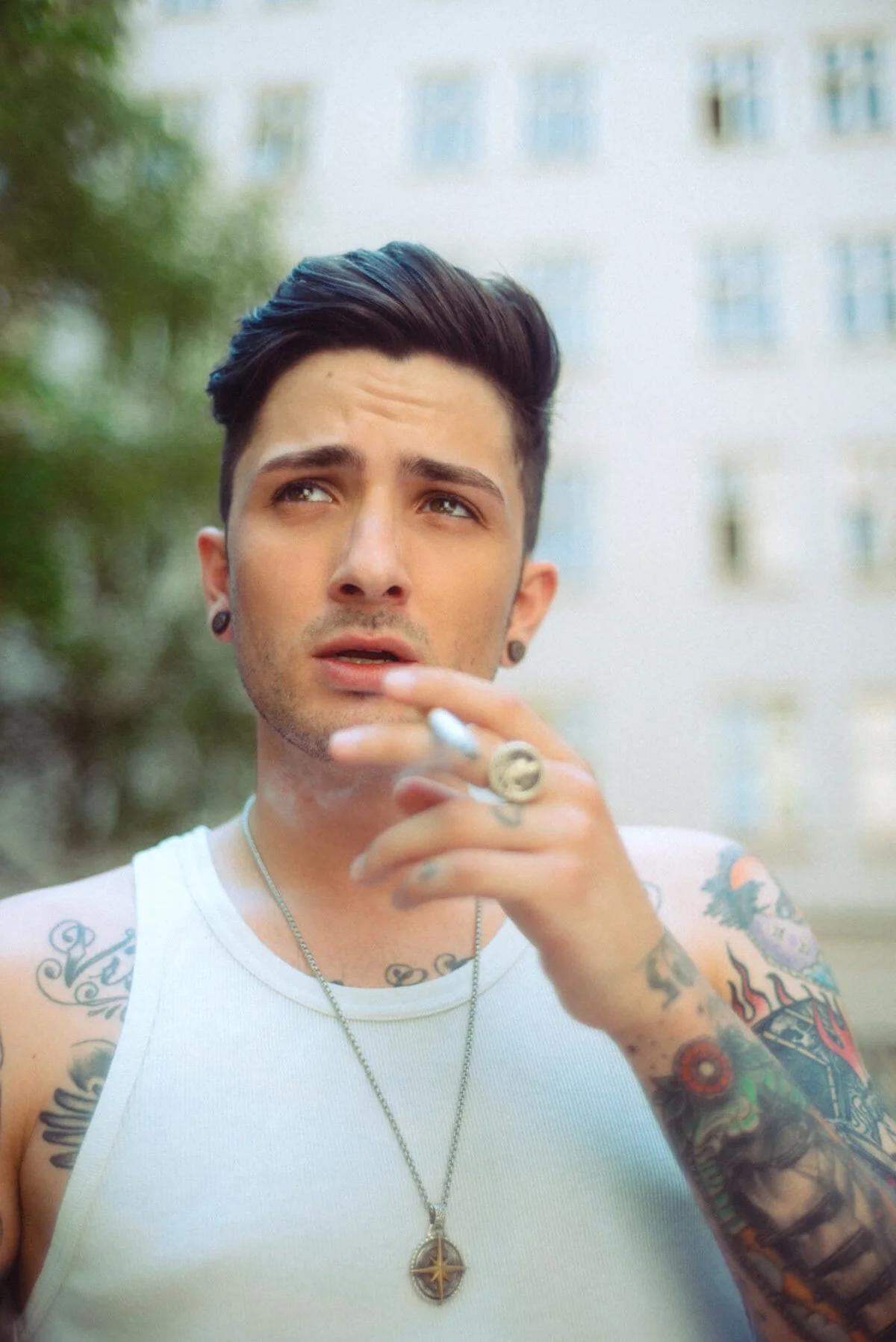 Portrait von Benjamin Mittelstädt bekannt aus "Berlin Tag und Nacht". Mit Tattoos, Piercings und einem weißen Tanktop, der draußen raucht, vor einem unscharfen Hintergrund mit Gebäuden und Bäumen.