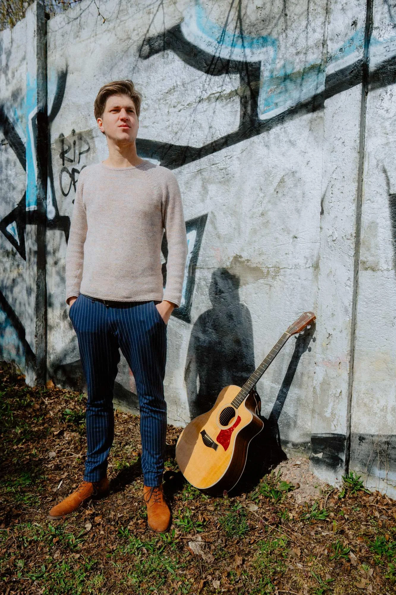 Falk Plücker steht vor einer Graffiti-Wand, trägt einen beigen Pullover, blaue kariertes Hosen und braune Stiefel. Er hat die Hände in den Taschen.