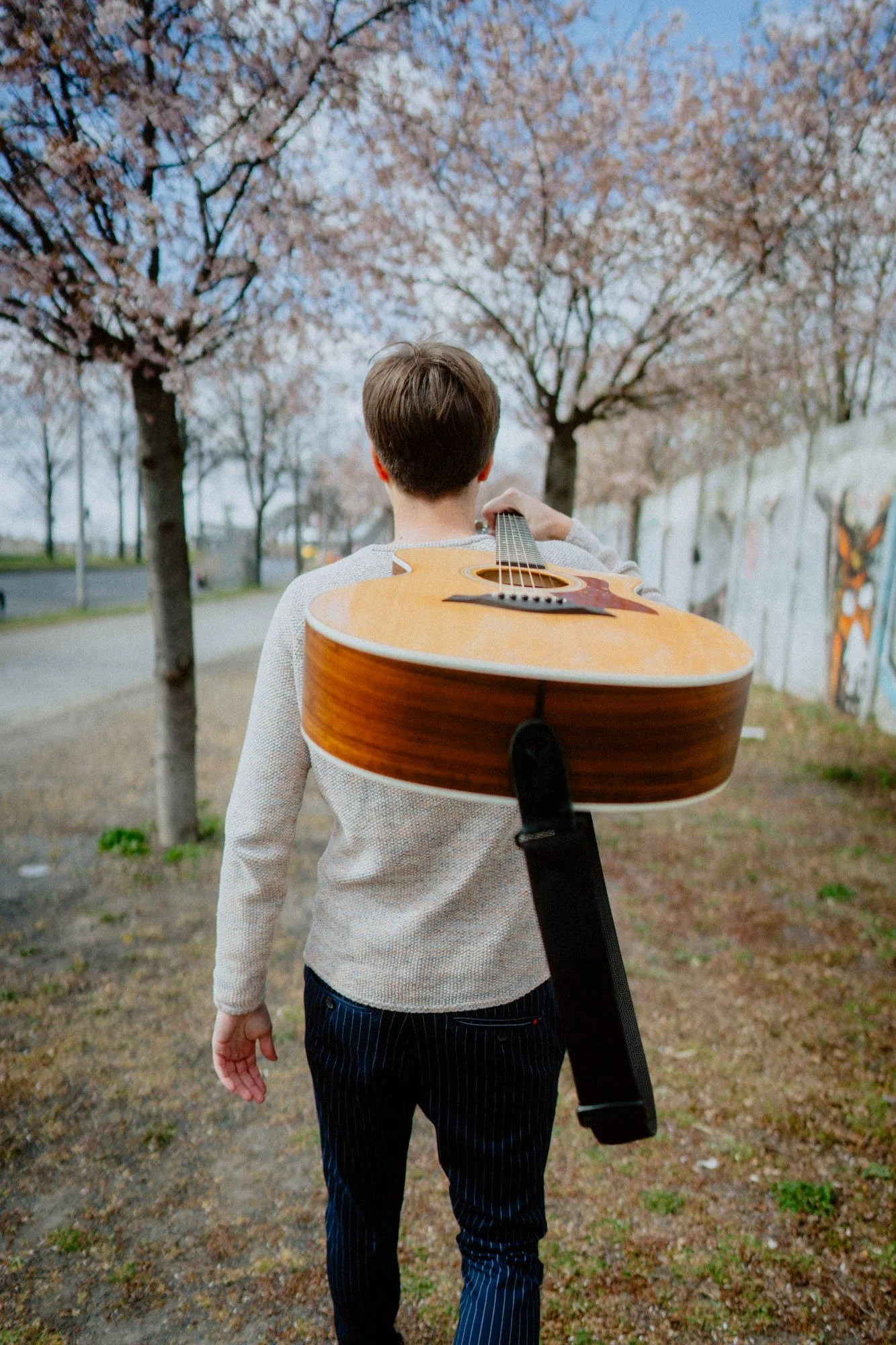 Liedermacher Falk mit Akustikgitarre auf dem Rücken, geht an blühenden Kirschbäumen entlang.