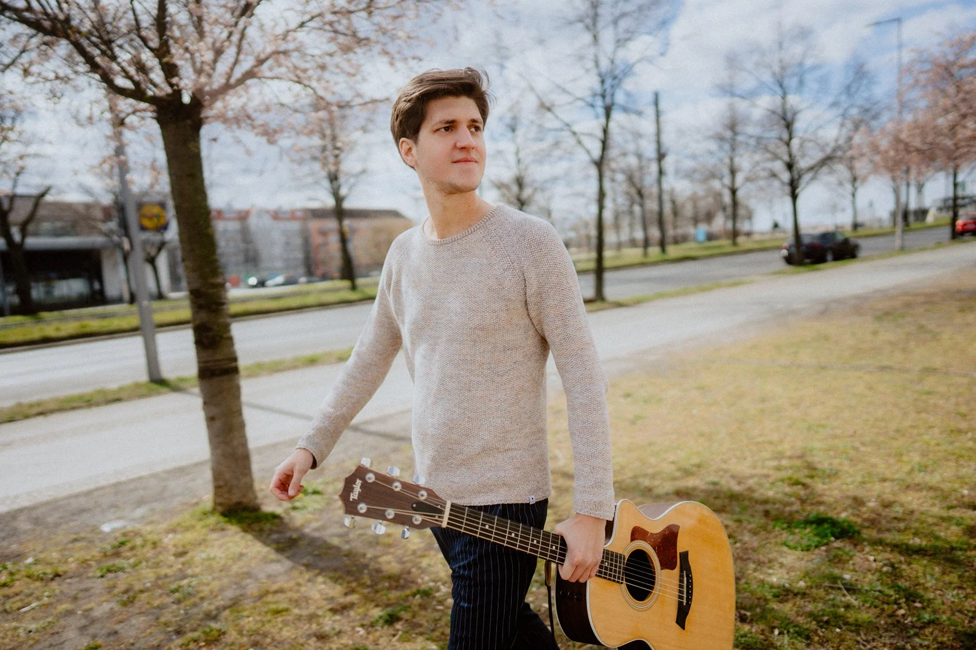 Falk Plücker mit braunen Haaren trägt ein hellgraues Pullover und hält eine Akustikgitarre in der Hand, während er durch eine parkähnliche Gegend mit Bäumen und Straßen geht. Es ist ein bewölkter Tag im Frühling.