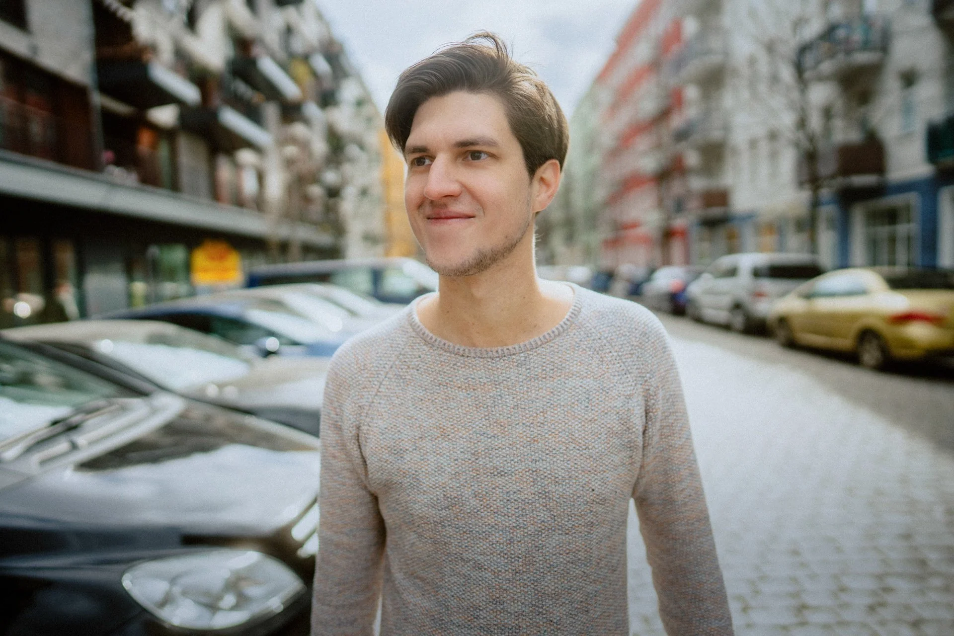 Liedermacher Falk aus Berlin in einem grauen Pullover auf einer Straße mit Parkplätzen und mehrstöckigen Gebäuden im Hintergrund.