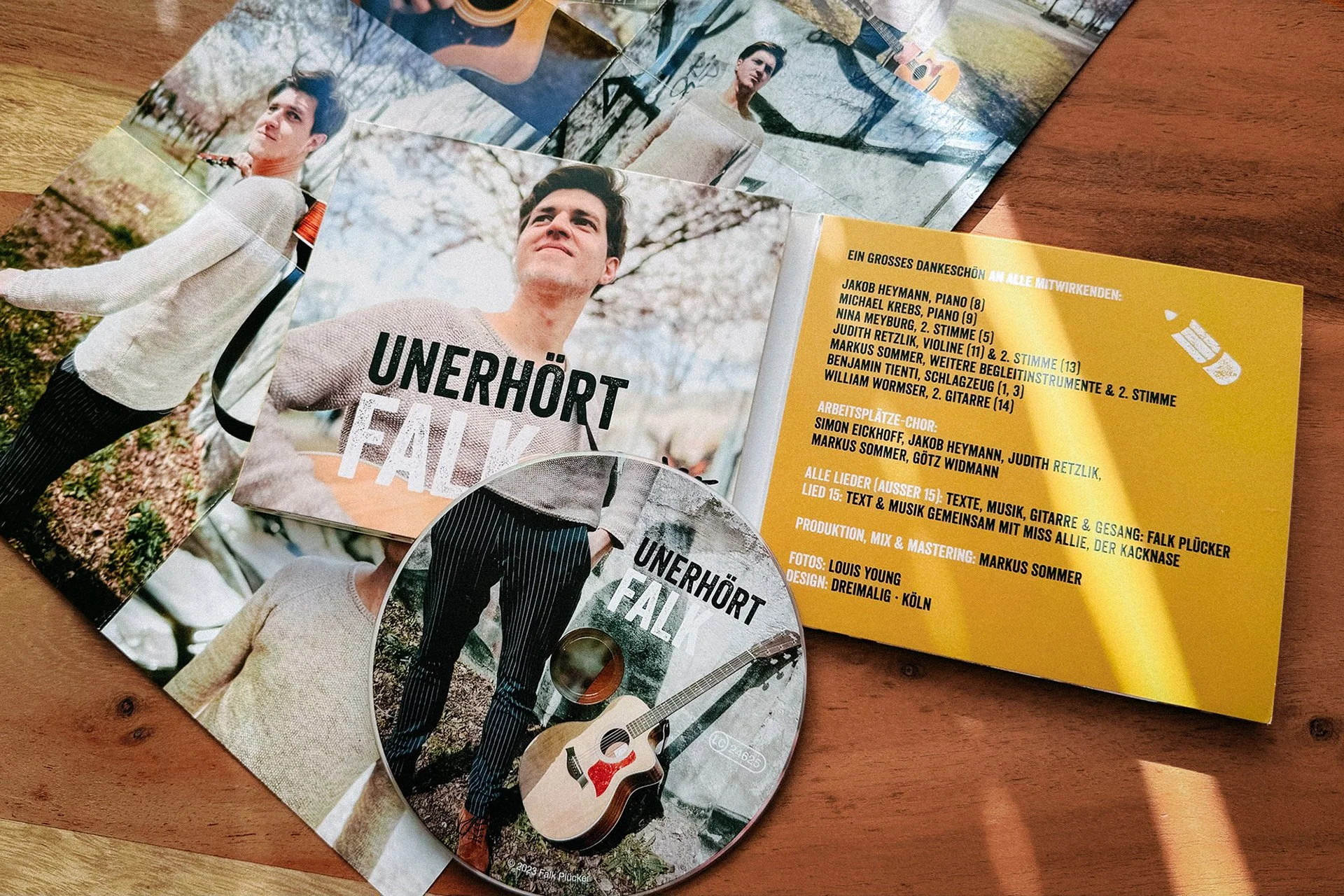 CD, Tüte und Flyer mit einem Mann im Freien, der ein Hauskleid trägt und eine Gitarre hält, sowie Textinformationen zum Konzert, darunter Liedtexte, Mitwirkende und Produktion