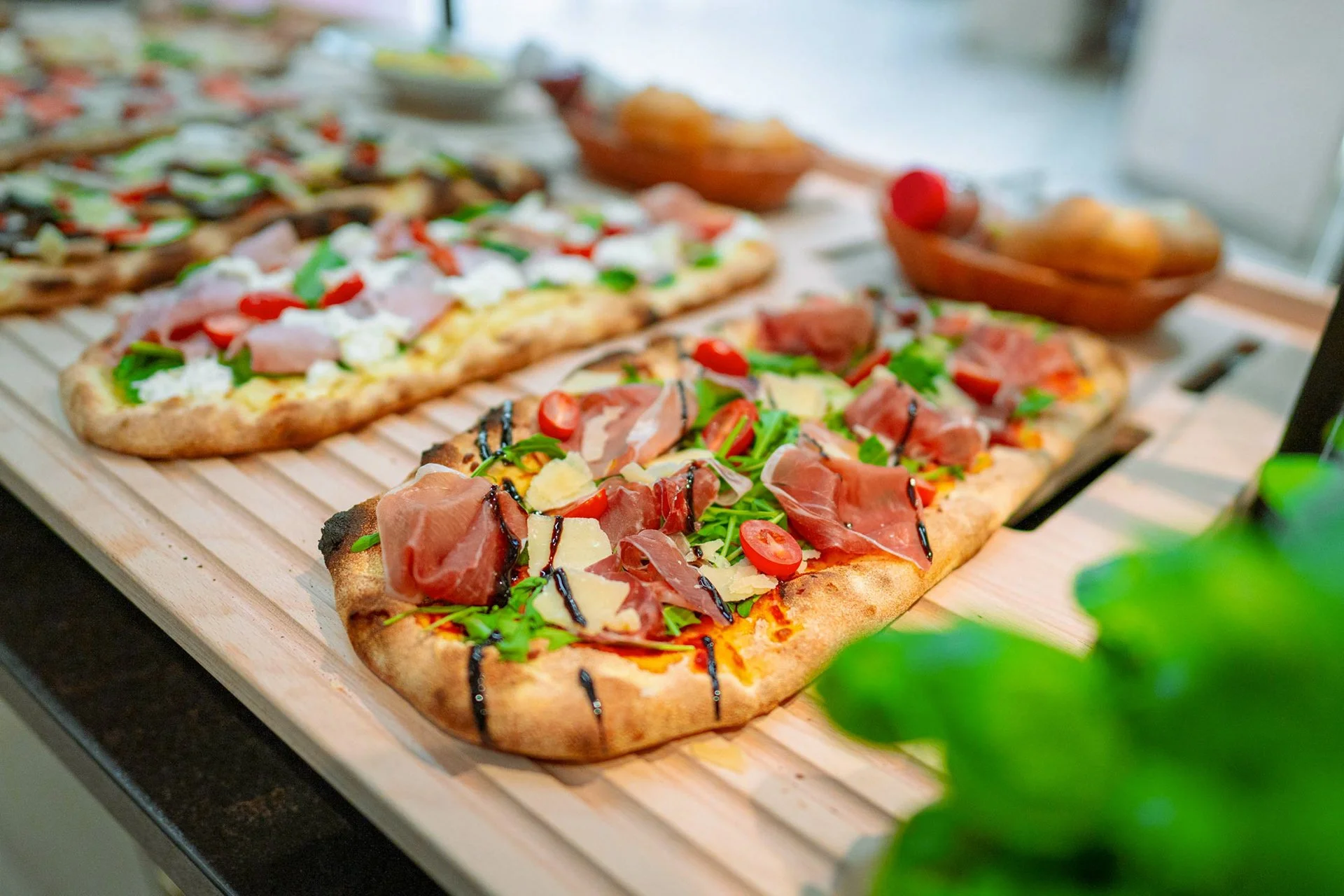 Verschiedene frisch gebackene Pizzen mit Belägen wie Schinken, Rucola, Tomaten, Käse und Balsamico-Glasur auf Holztablett