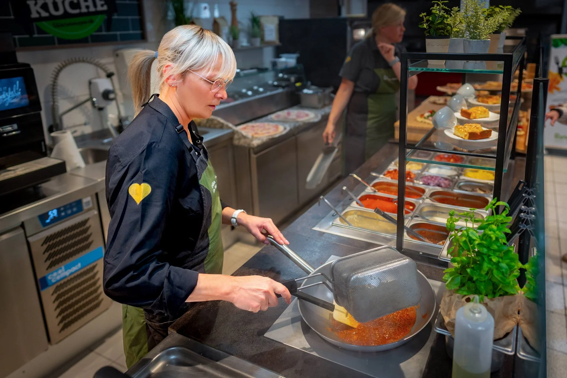 Eine Frau kocht auf einer Gastronomiestation, während eine andere Person im Hintergrund arbeitet. Auf der Theke sind verschiedene Saucen und Toppings in Behältern. Die Frau in Vordergrund trägt eine schwarze Jacke und eine grüne Schürze.