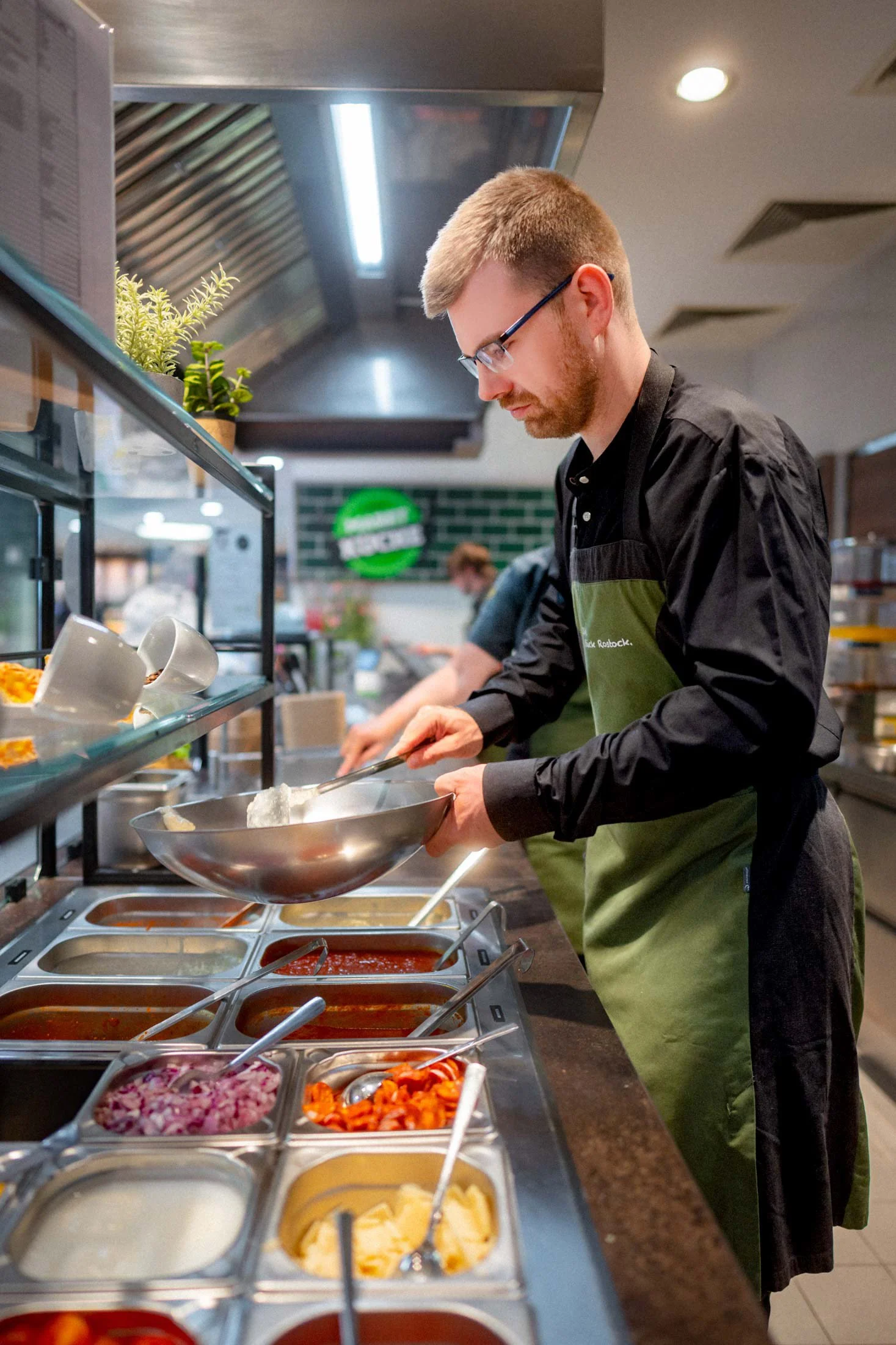 Ein männlicher Mitarbeiter bereitet Speisen an einer Selbstbedienungs-Theke in einem Restaurant zu. Im Hintergrund sind weitere Mitarbeiter sichtbar.