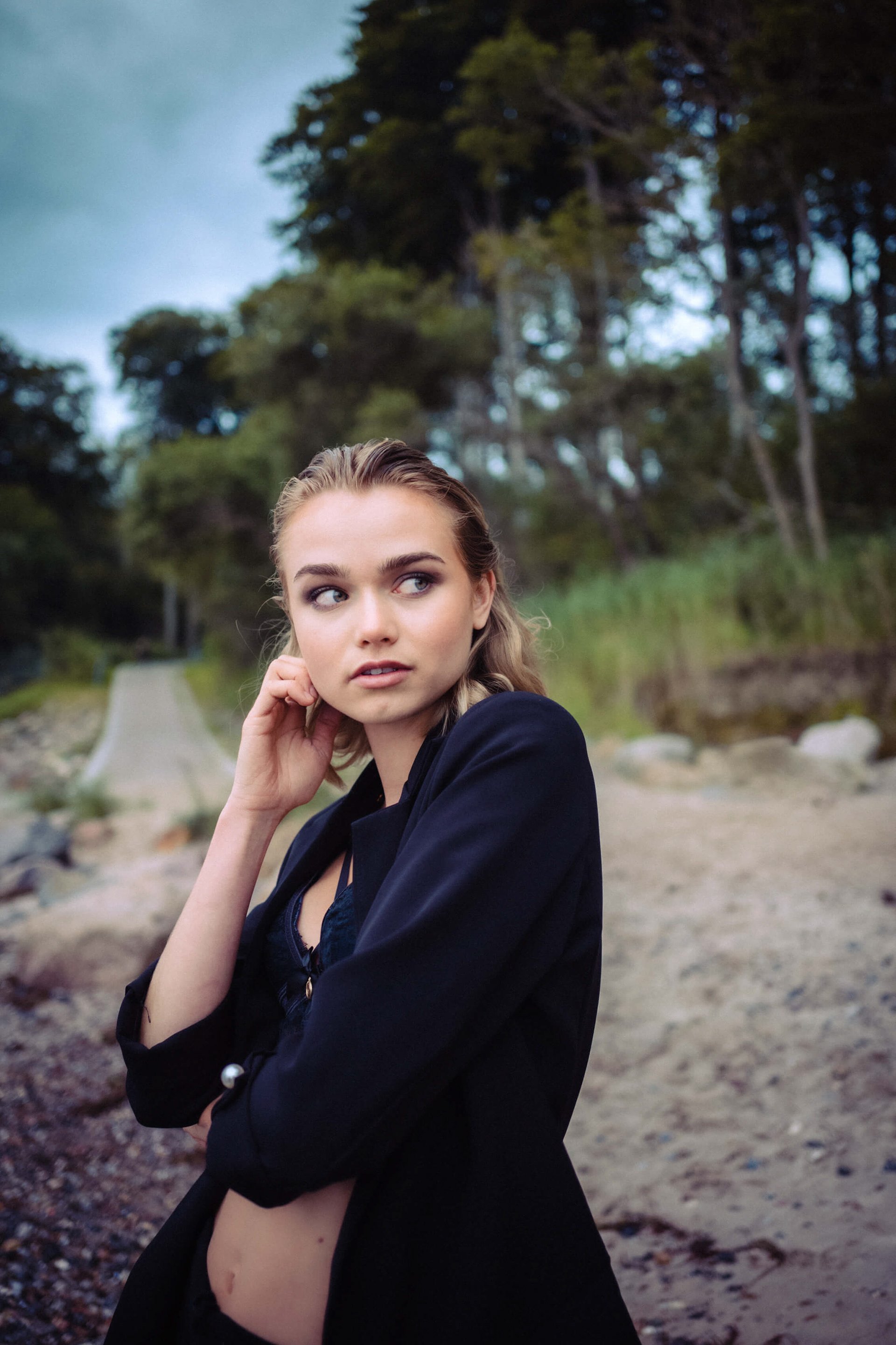 Lilian Franz am Strand, trägt schwarze Jacke, schaut seitlich, blondes Haar, natürliche Umgebung.