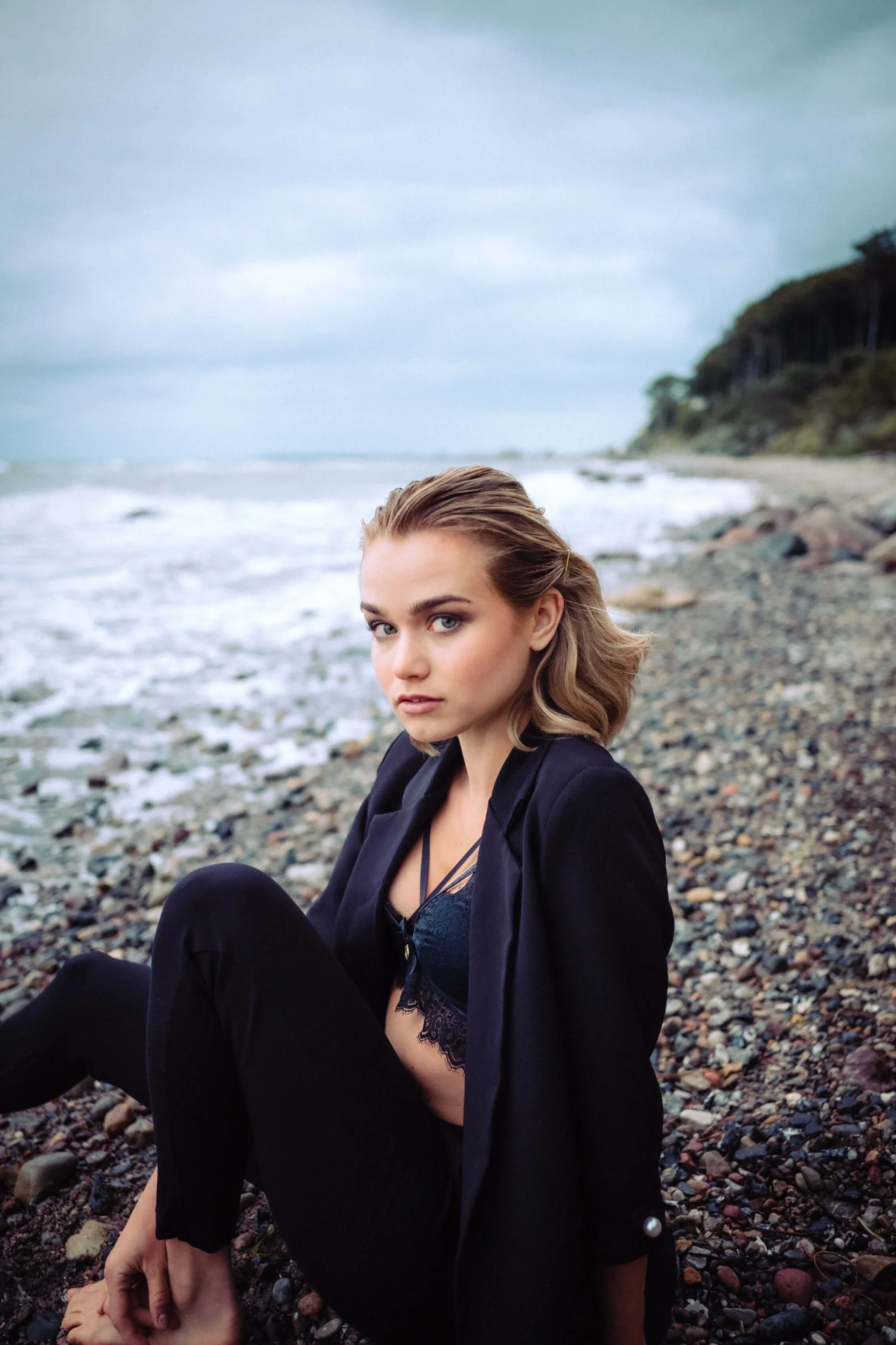 Lilian Franz am Strand bei bewölktem Himmel, sitzend auf Kieselsteinen, trägt schwarze Kleidung und blickt in die Kamera.