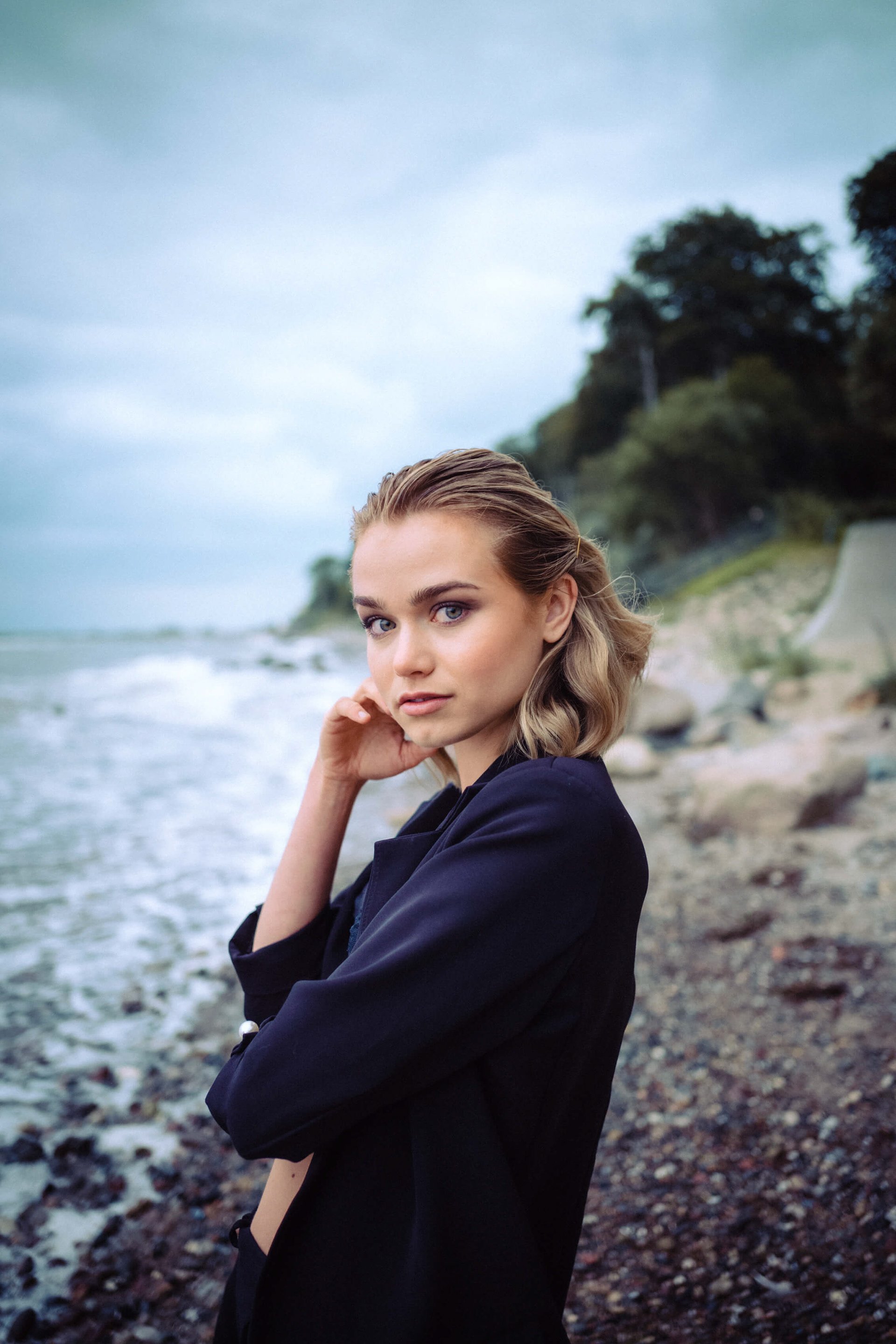 Junge Frau am Strand bei bewölktem Himmel, trägt dunkle Jacke, blickt in die Kamera, im Hintergrund Meer und Bäume.