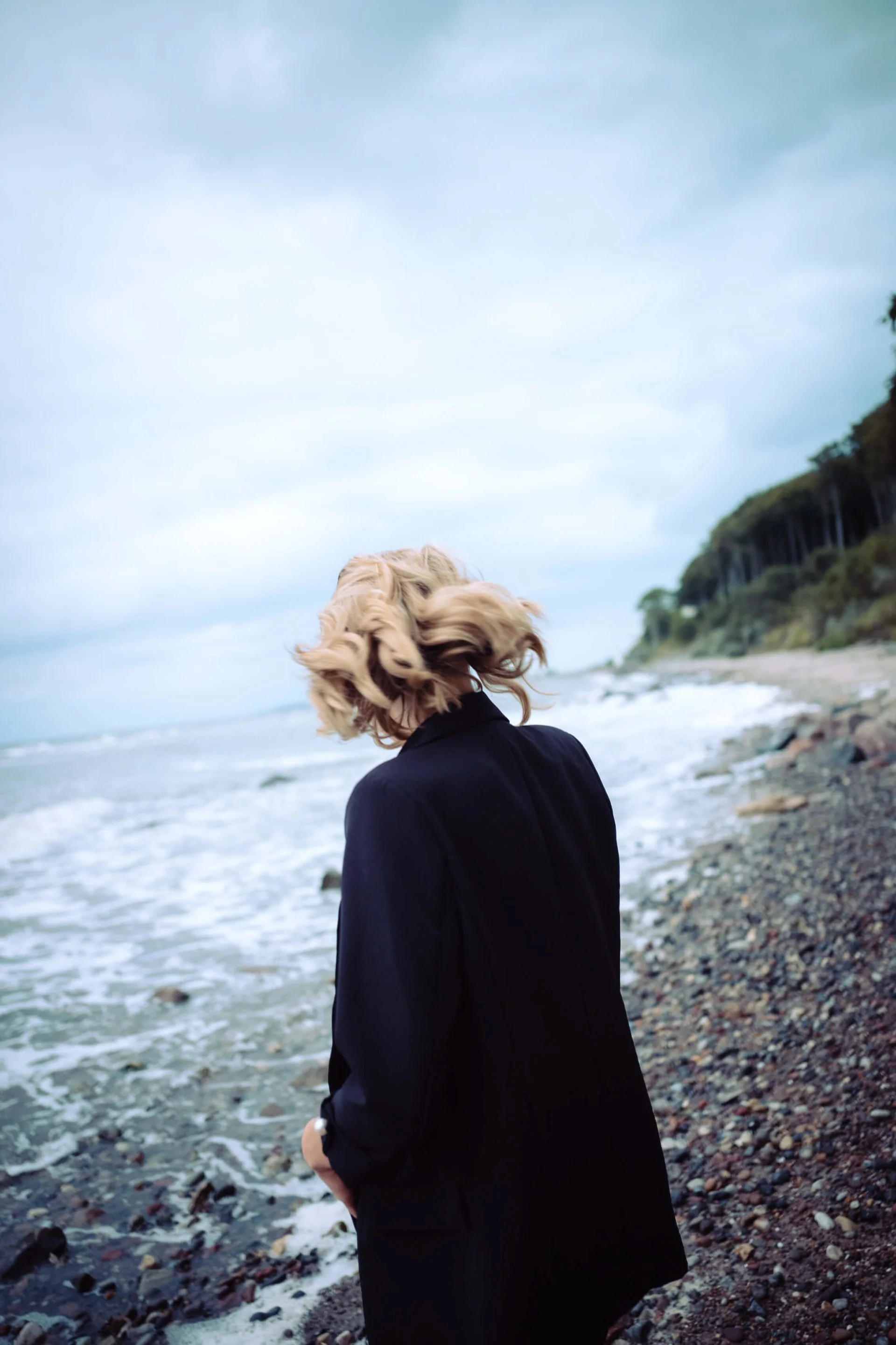 Lilian Franz mit blondem, lockigem Haar trägt schwarze Jacke und steht am Strand, blickt aufs Meer, im Hintergrund bewölkter Himmel und grüne Klippen.