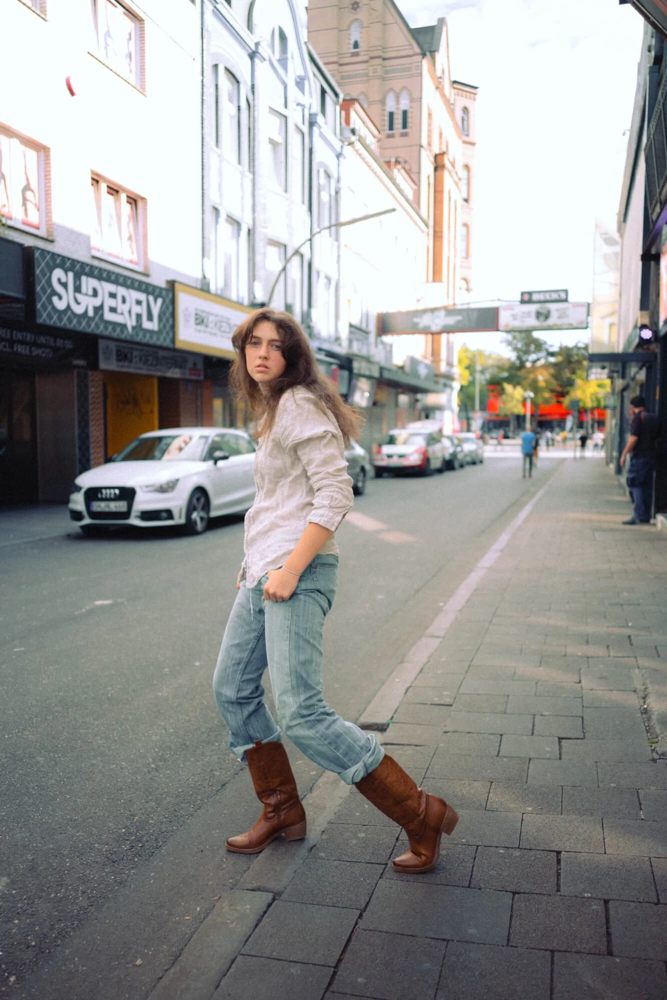 Carlotta Josefine Pahl mit braunen, lockigen Haaren trägt eine bequeme weiße Bluse, blaue Jeans und braune Stiefel. Sie steht auf einem Gehweg in einer belebten Stadtstraße mit Geschäften und parkenden Autos im Hintergrund.