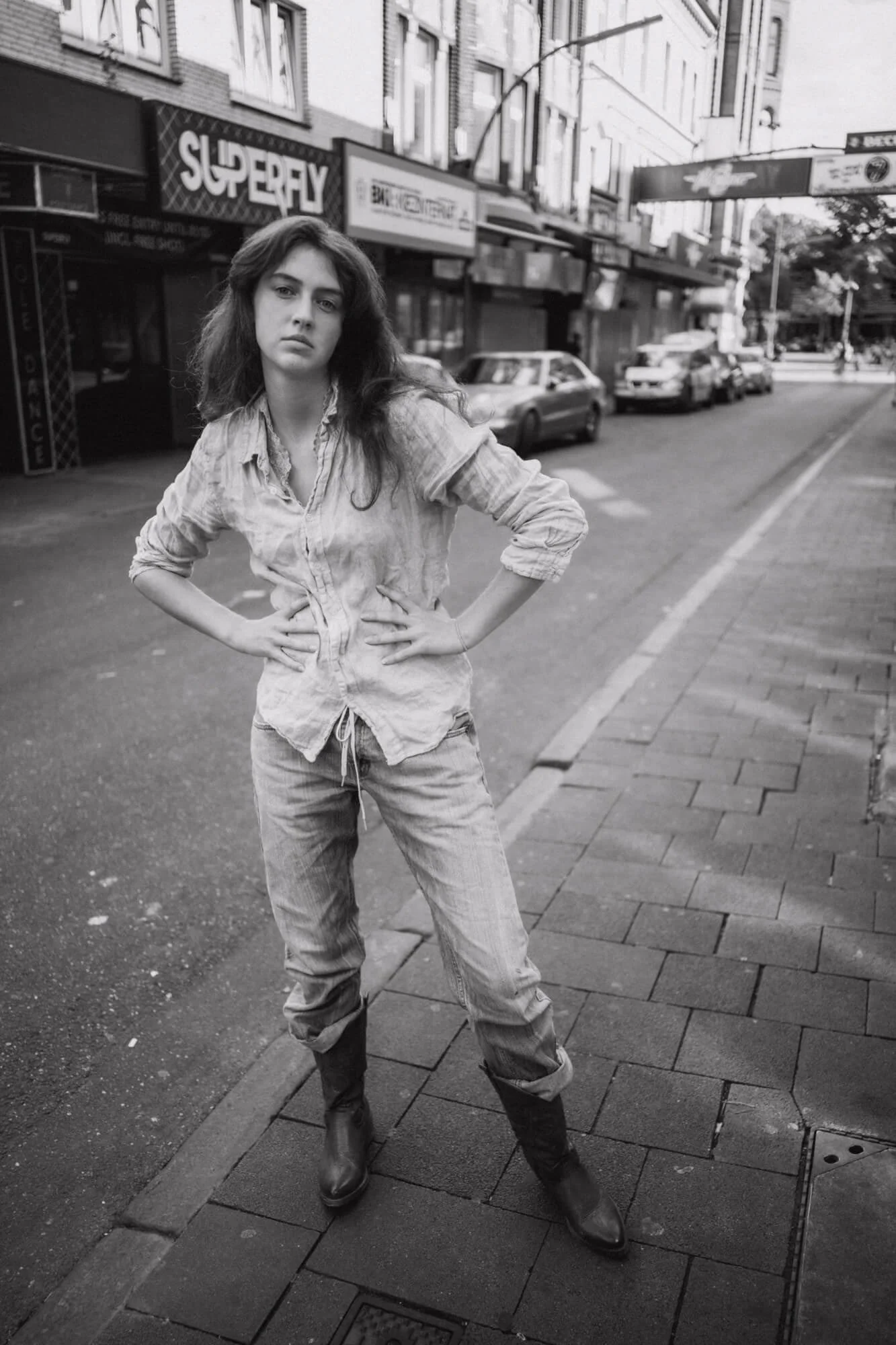 Schwarz-weiß-Foto von Carlotta Josefine Pahl  mit lässigem Kleid, stehende Pose mit Händen in den Hüften, auf Gehweg in einer Stadtstraße, im Hintergrund Autos und Geschäfte.