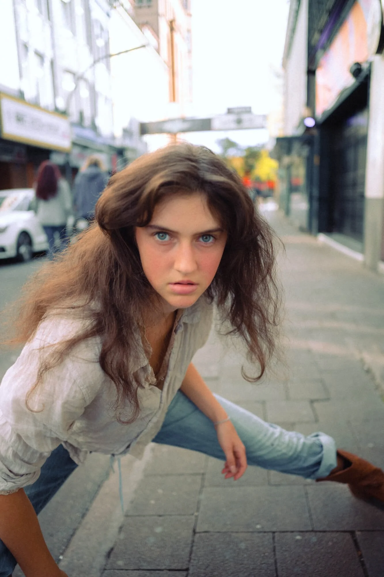 Carlotta Josefine Pahl, die auf der Straße in die Kamera schaut, im Vordergrund kniende Haltung, graues Oberteil, blaue Augen, braune, lockere Haare, städtische Umgebung mit Geschäften und Autos im Hintergrund.