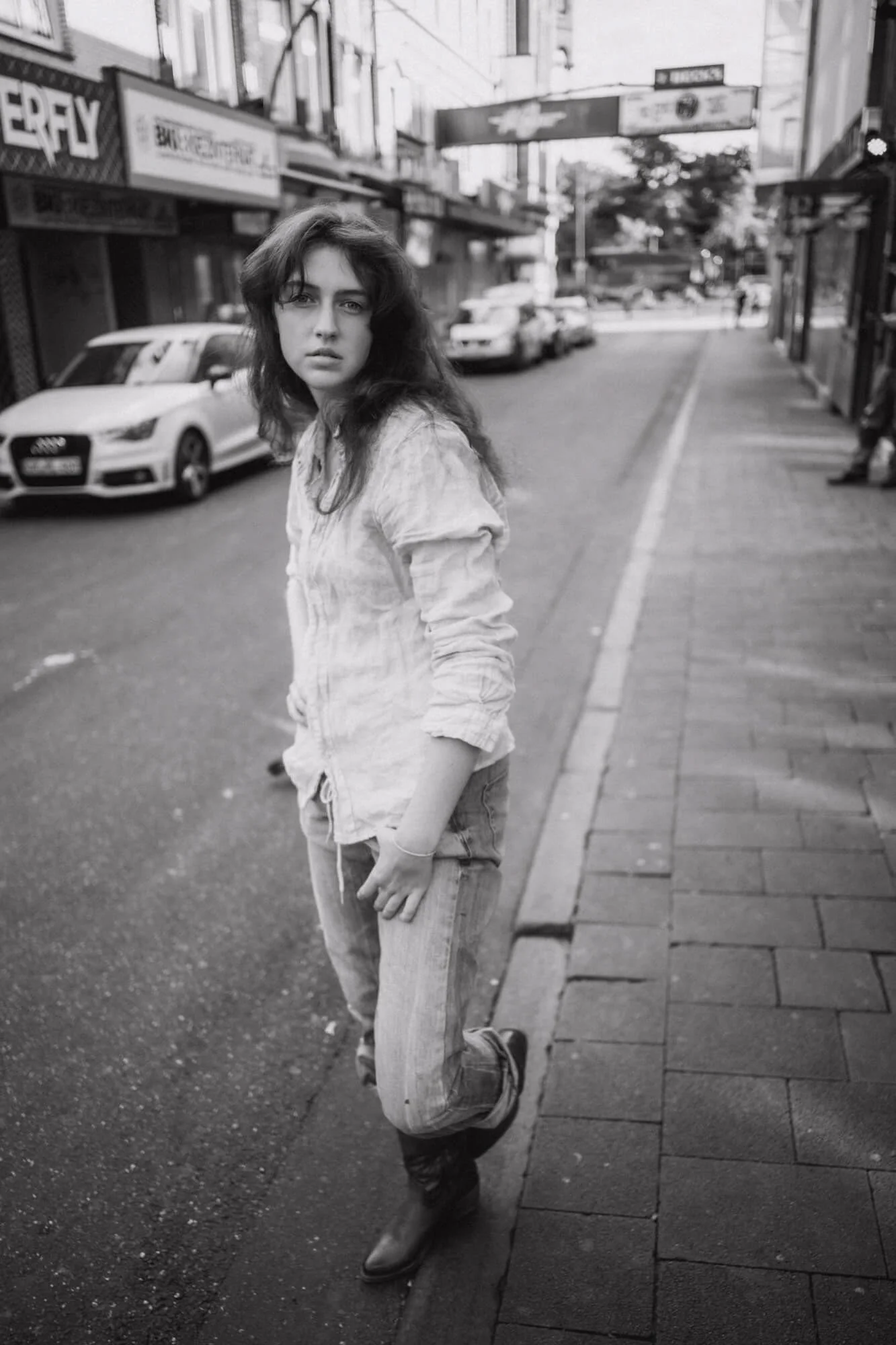 Schwarz-weiß-Foto von Carlotta Josefine Pahl, die an einer Straßenseite steht, mit dunklem, gewelltem Haar, legerer Kleidung und Boots, in einer städtischen Umgebung mit Autos und Geschäften im Hintergrund.