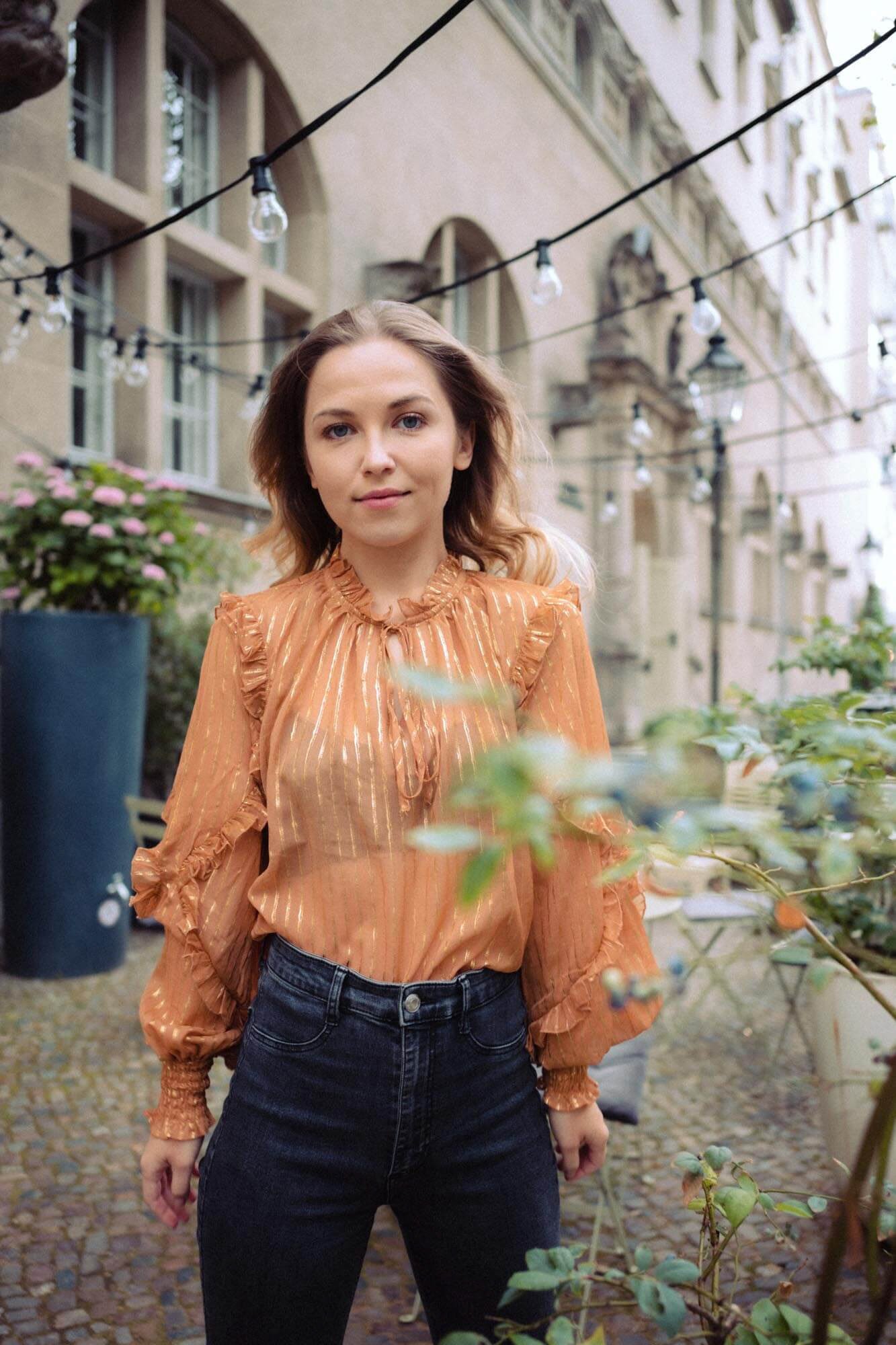 Marija Mauer mit braunen, gewellten Haaren trägt eine orange Rüschenbluse und dunkle Jeans, steht vor einem Gebäude im Freien, mit Lichterketten über ihr, und Pflanzen im Vordergrund.