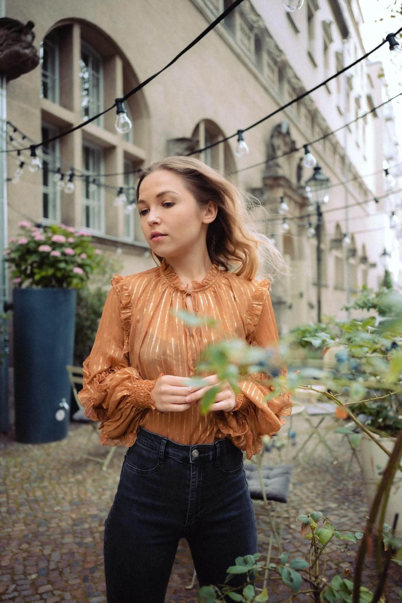 Marija Mauer mit braunen, gewellten Haaren steht im Freien in einem urbanen Garten mit Pflanzkübeln und bewölktem Himmel, umgeben von alten Gebäuden und Lichterketten.