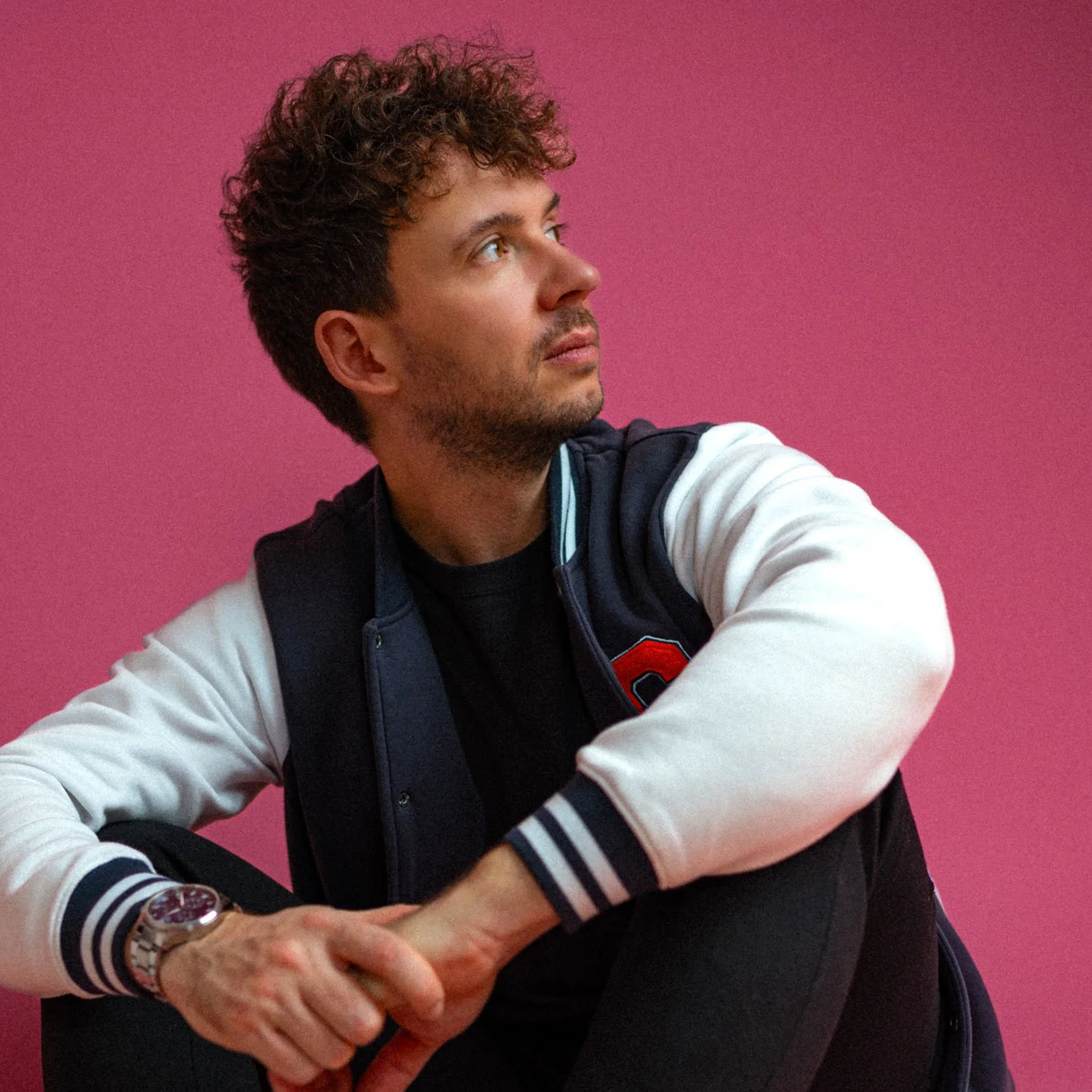 Junge Mann mit braunen, lockigen Haaren und einem Bart sitzt vor pinkem Hintergrund, trägt eine schwarze Jacke mit weißen Ärmeln, ein schwarzes T-Shirt und einen goldenen Armband.