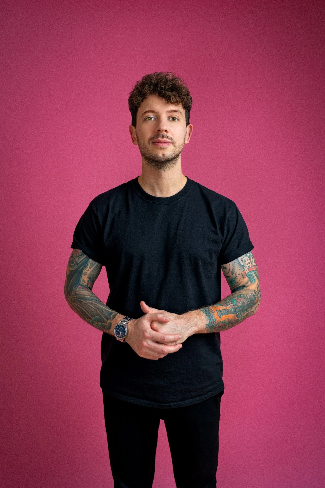 Ein junger Mann mit Tattoos, schwarzen T-Shirt und Uhr steht vor einem pinken Hintergrund.