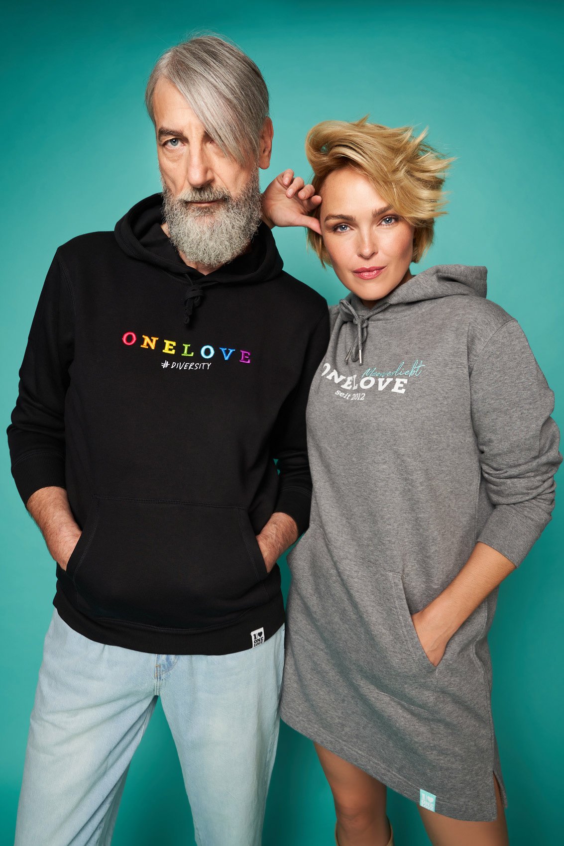 Zwei Menschen stehen vor einem grünen Hintergrund, tragen Hoodies mit bunten Schriftzügen, der Mann hat einen grauen Bart und das Haar, die Frau kurze blonde Haare.