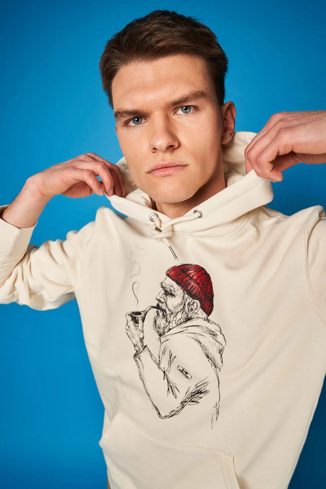 Junger Mann in cremfarbener Kapuzenjacke mit Collage von Skizze eines Mannes mit roter Mütze und Pfeife auf Brust. Hintergrund blau.