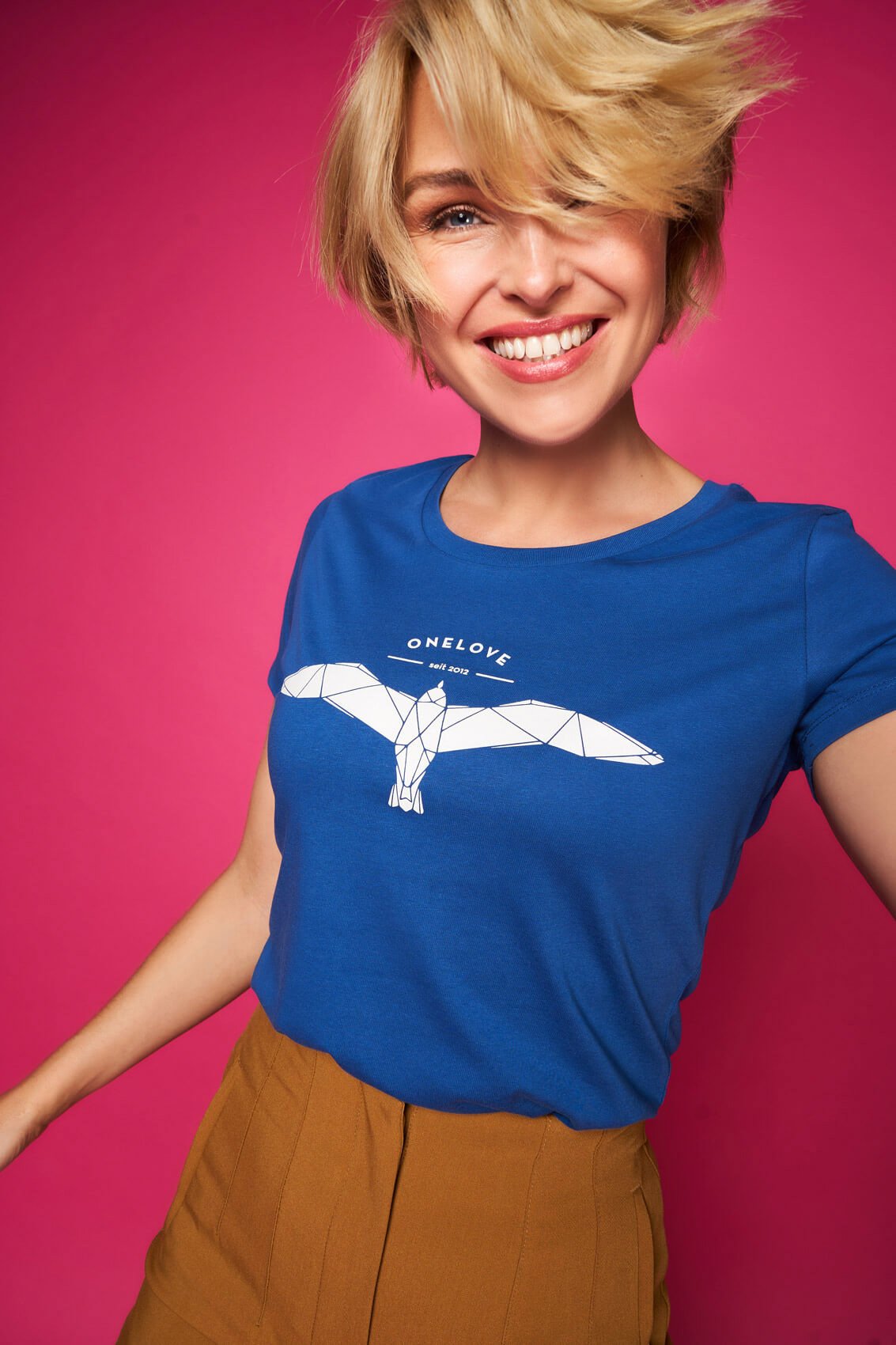 Junge Frau mit blondem, kurzen, zerzaustem Haar, lachend vor einem pinken Hintergrund, trägt ein blaues T-Shirt mit einem weißen Vogelmotiv und dem Text 'ONELOVE seit 2012', dazu braune Hosen.