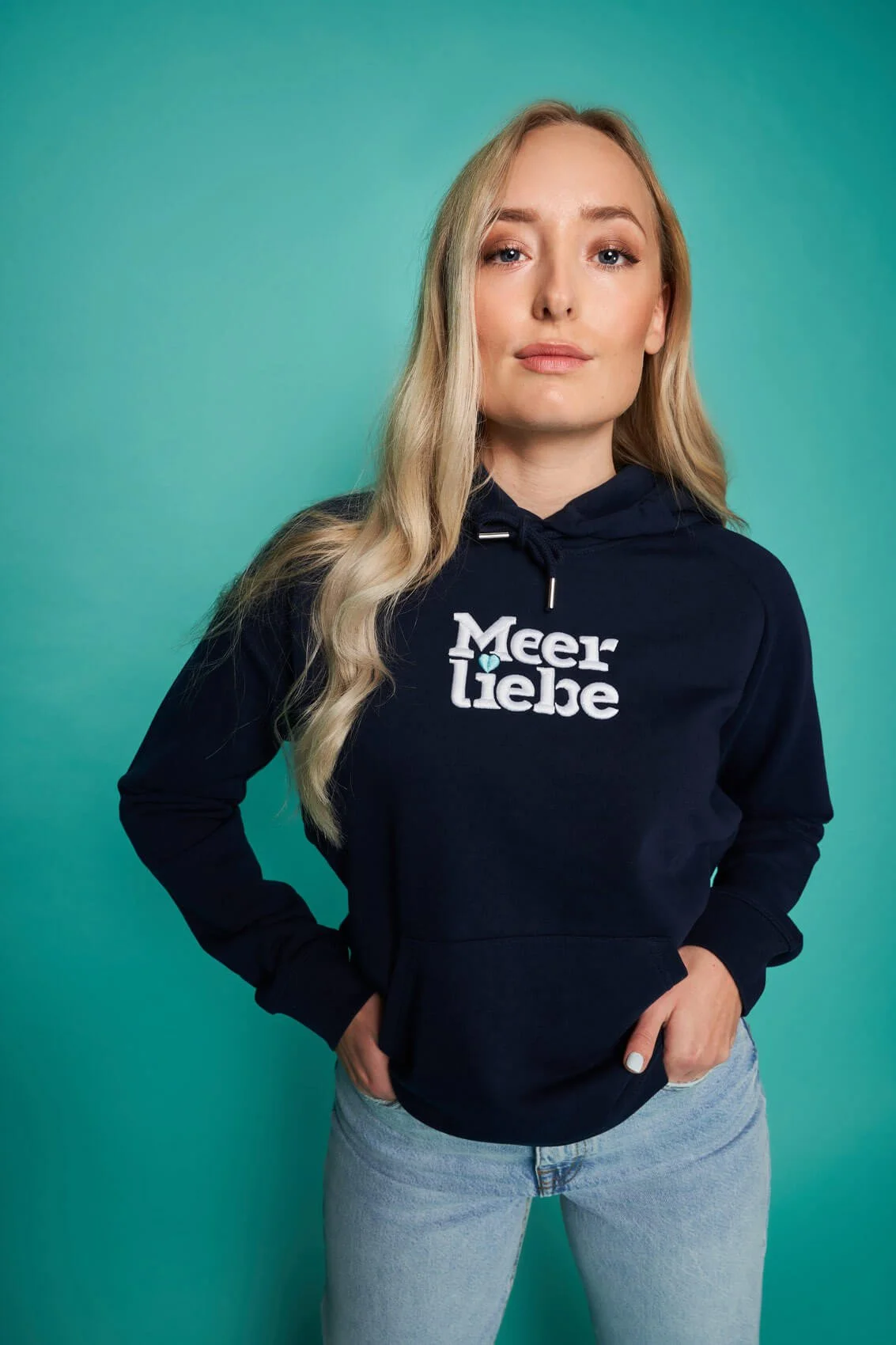 Junge Frau mit blonden Haaren trägt eine schwarze Hoodie mit der Aufschrift "Meer Liebe" und steht vor einem grünen Hintergrund.