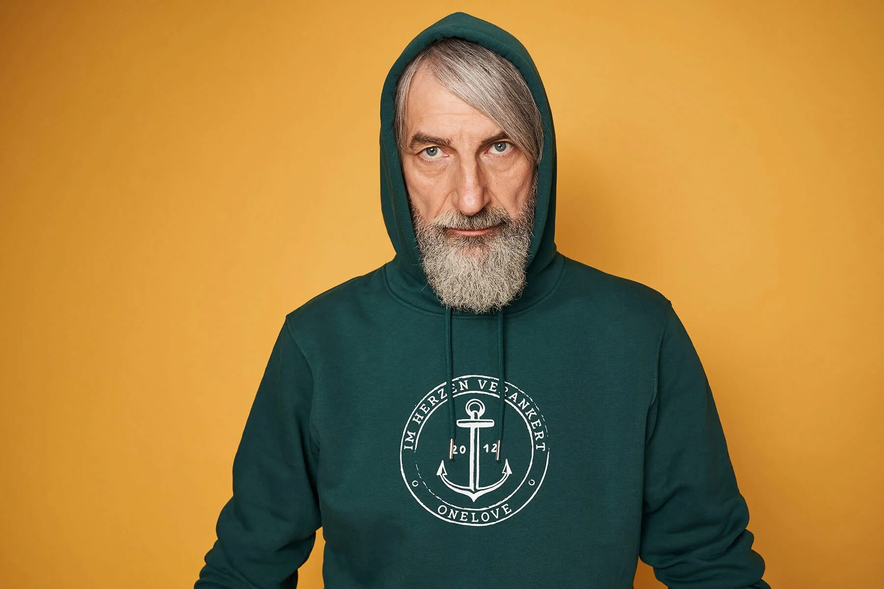 Ein älterer Mann mit grauem Bart und grauen Haaren trägt einen dunkelgrünen Hoodie mit Kapuze vor einem gelben Hintergrund.