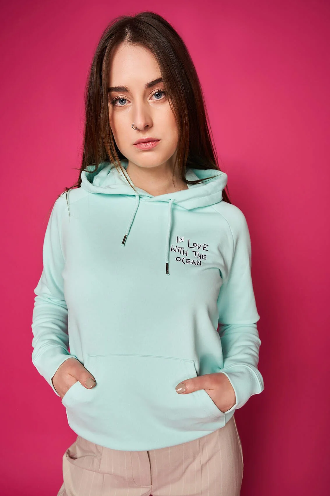 Junge Frau mit braunen Haaren, in einem hellblauen Hoodie mit Kapuze, die Hände in den Kängurutaschen, vor pinkem Hintergrund.