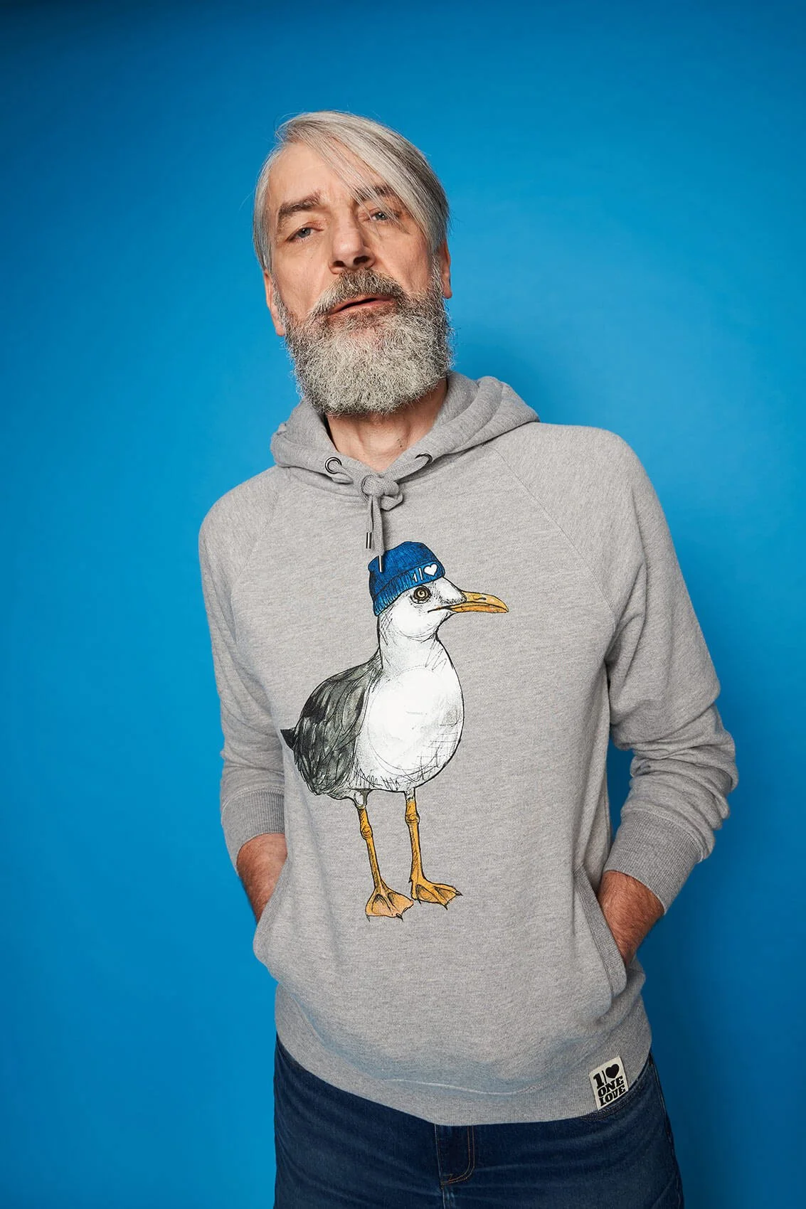 Ein älterer Mann mit grauem Bart trägt einen grauen Hoodie mit einer Zeichnung eines Möwenkopfs, der eine blaue Mütze trägt, gegen einen blauen Hintergrund.