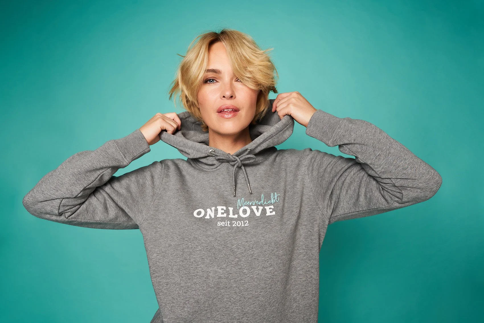 Junge Frau trägt grauen Hoodie mit Aufschrift 'ONE LOVE seit 2012' vor einfarbigem Hintergrund.