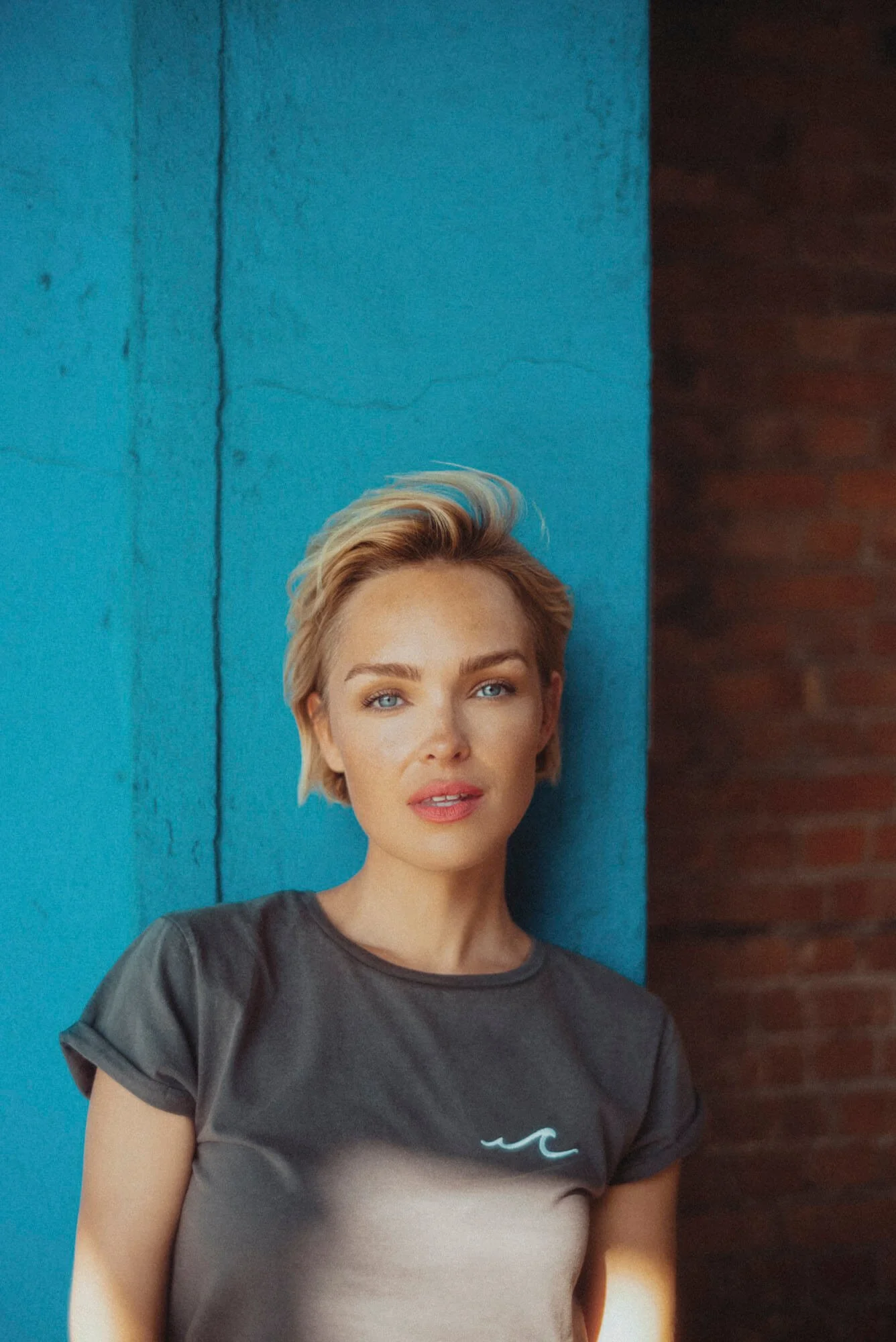 Junge Frau mit kurzen blonden Haaren und blauen Augen steht vor einer blauen Wand, trägt ein graues T-Shirt, Blick in die Kamera gerichtet.