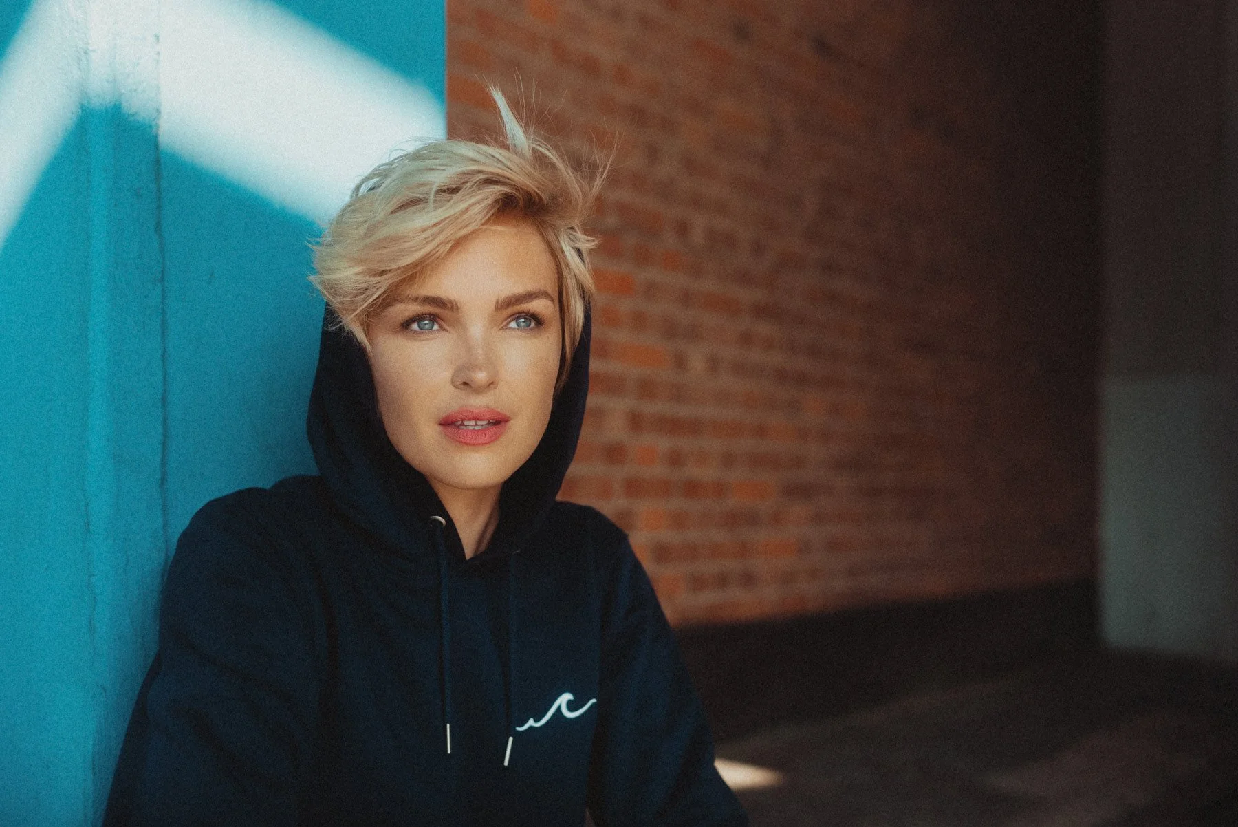 Junge Frau mit kurzen blondem Haar, trägt einen schwarzen Hoodie, sitzt an einer gelb-blauen Wand und blickt in die Ferne.