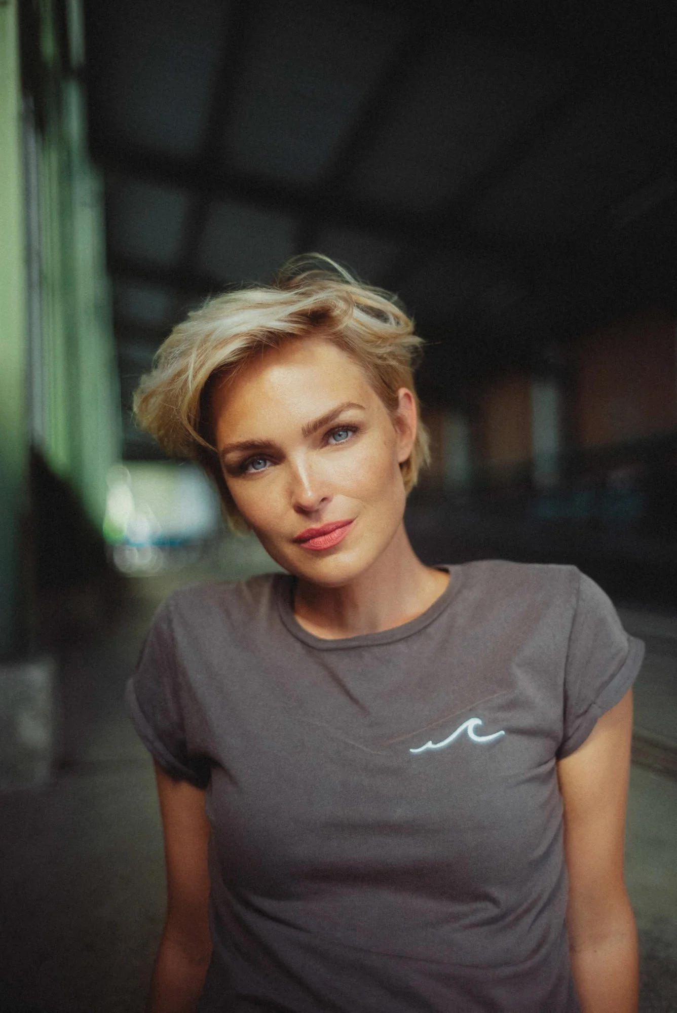 Ein älterer Blondschopf mit blauen Augen trägt ein graues T-Shirt mit einer kleinen Wellen-Logo. Sie steht auf einer Eisenbahnbrücke im Schatten, blickt in die Kamera, lächelt leicht.