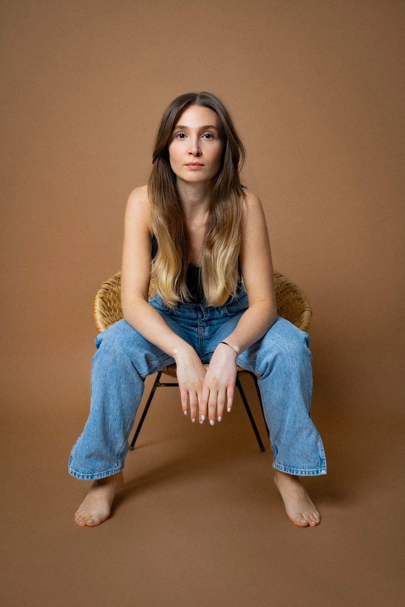 Schauspielerin Özge Kayalar sitzt barfuß auf einem Stuhl vor einem braunen Hintergrund.
