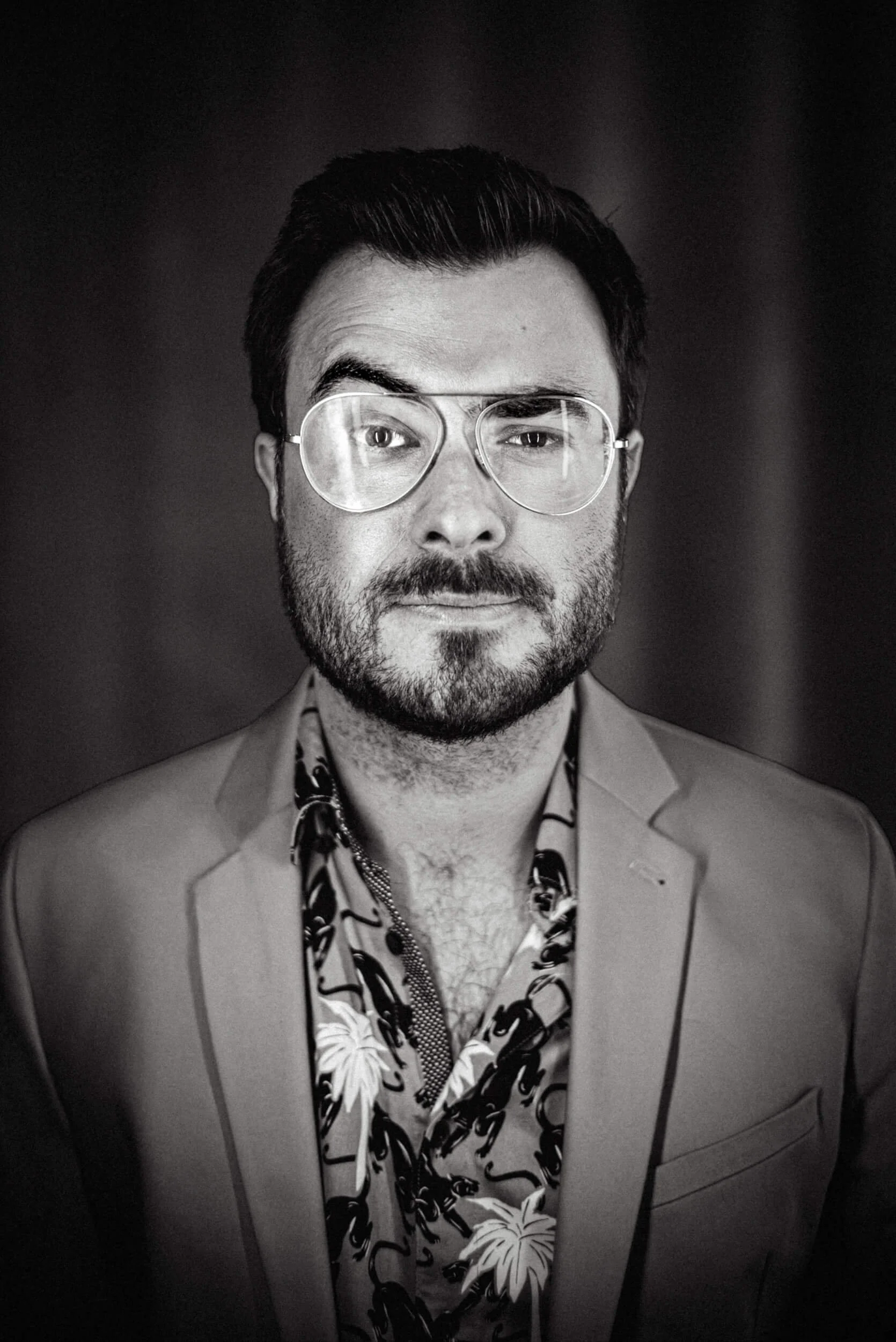 Schwarz-weiß-Portrait von Quiz-Master Sebastian Klussmann mit dunklem Haar, Brille, Vollbart, blumigem Hemd und Anzugjacke vor dunklem Hintergrund.