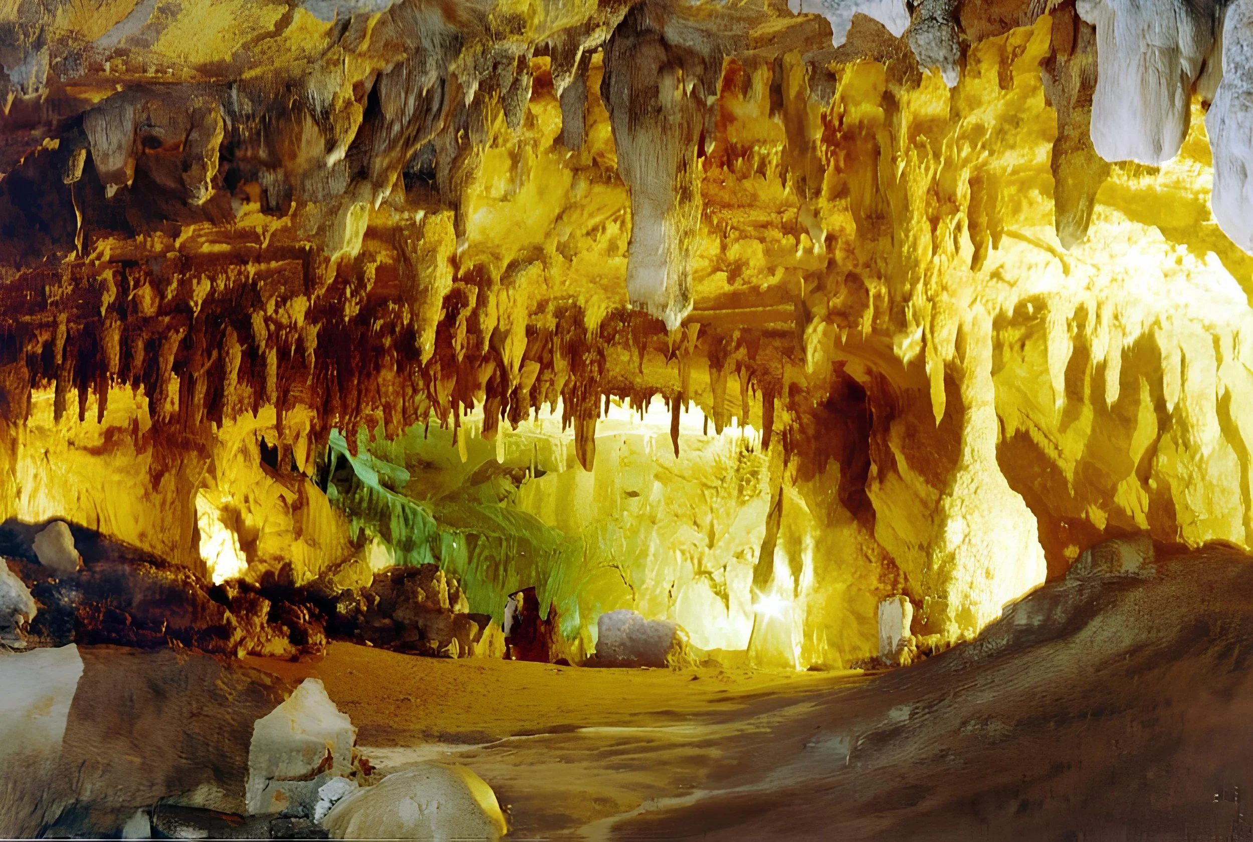 Inside the Lombrives cave with stalactites hanging from the ceiling, illuminated by yellow and green lighting, with a sandy floor and large rocks.