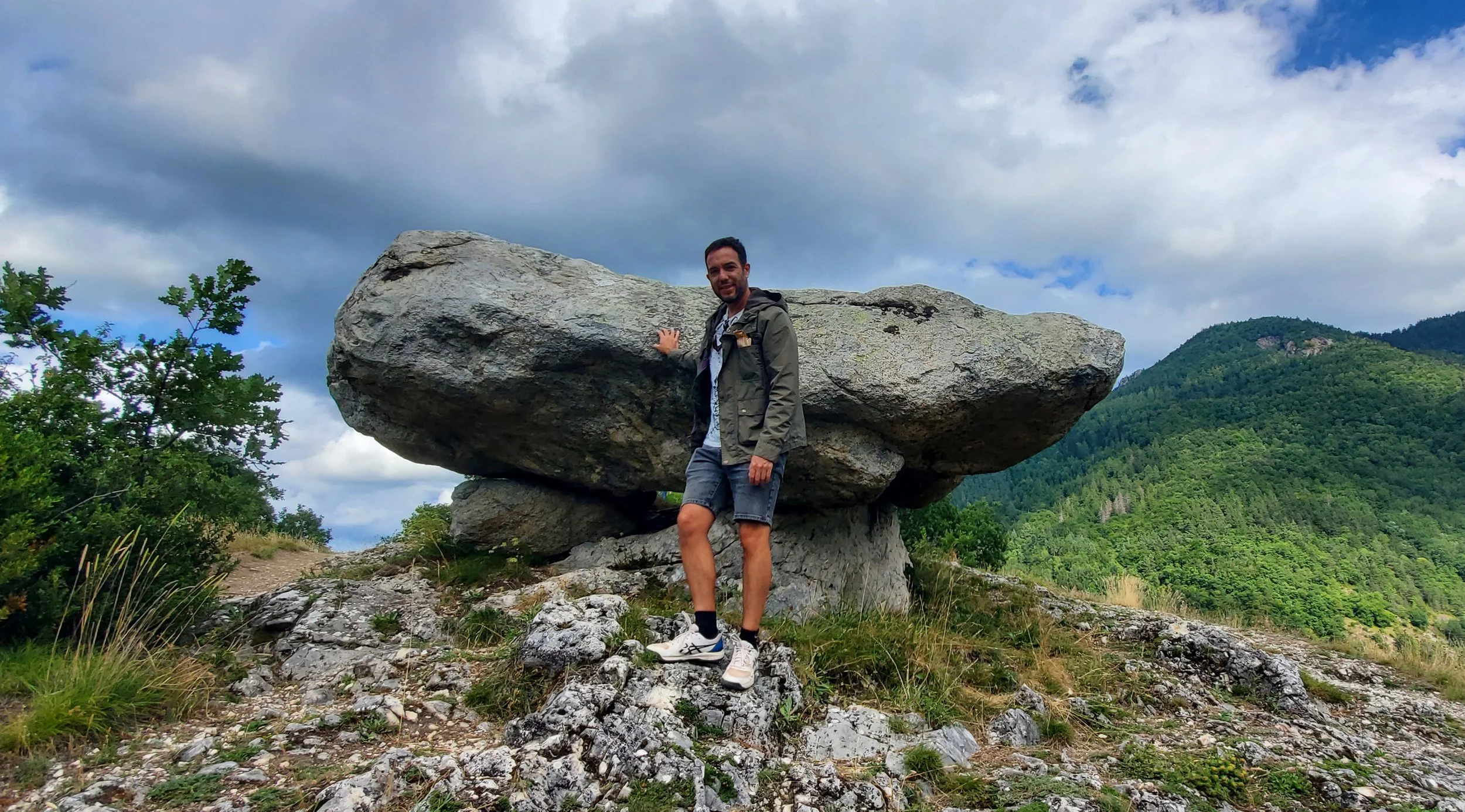 Arcturus standing in front of a large dolmen with mountains and green trees in the background, under a partly cloudy sky.