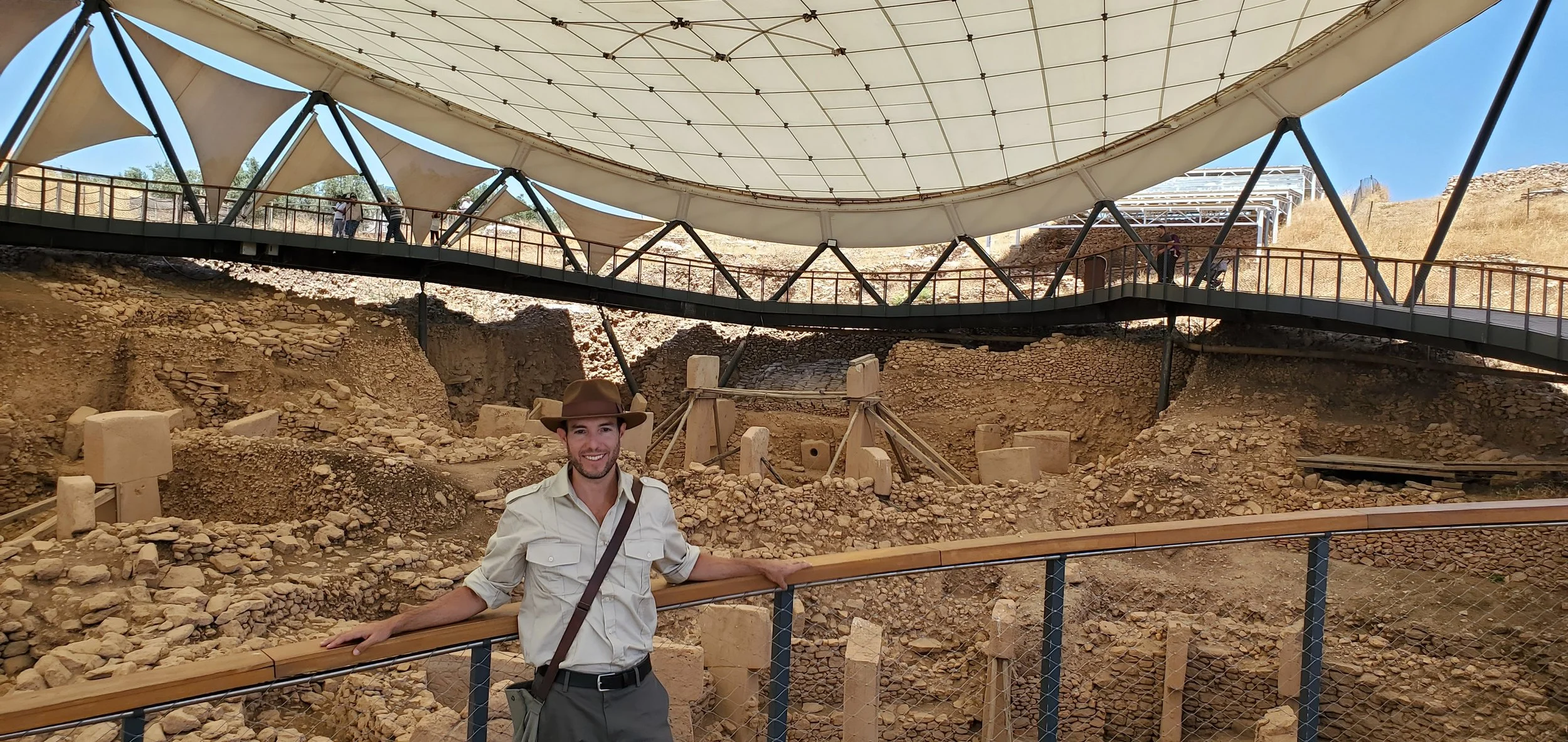 A smiling man in a beige safari hat and light shirt standing in Gobekli Tepe archaeological site under a large, modern canopy structure with walkways and a railing.