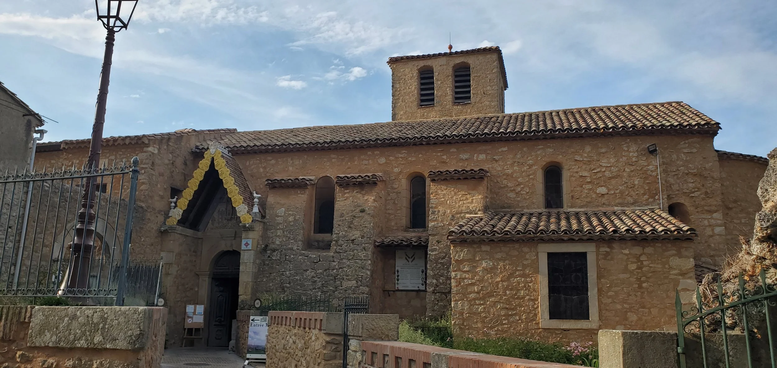 The old stone church of Mary Magdalene in Rennes-le-Chateau with a tiled roof, arched windows, and a small tower on top, surrounded by a gated fence and a stone pathway.