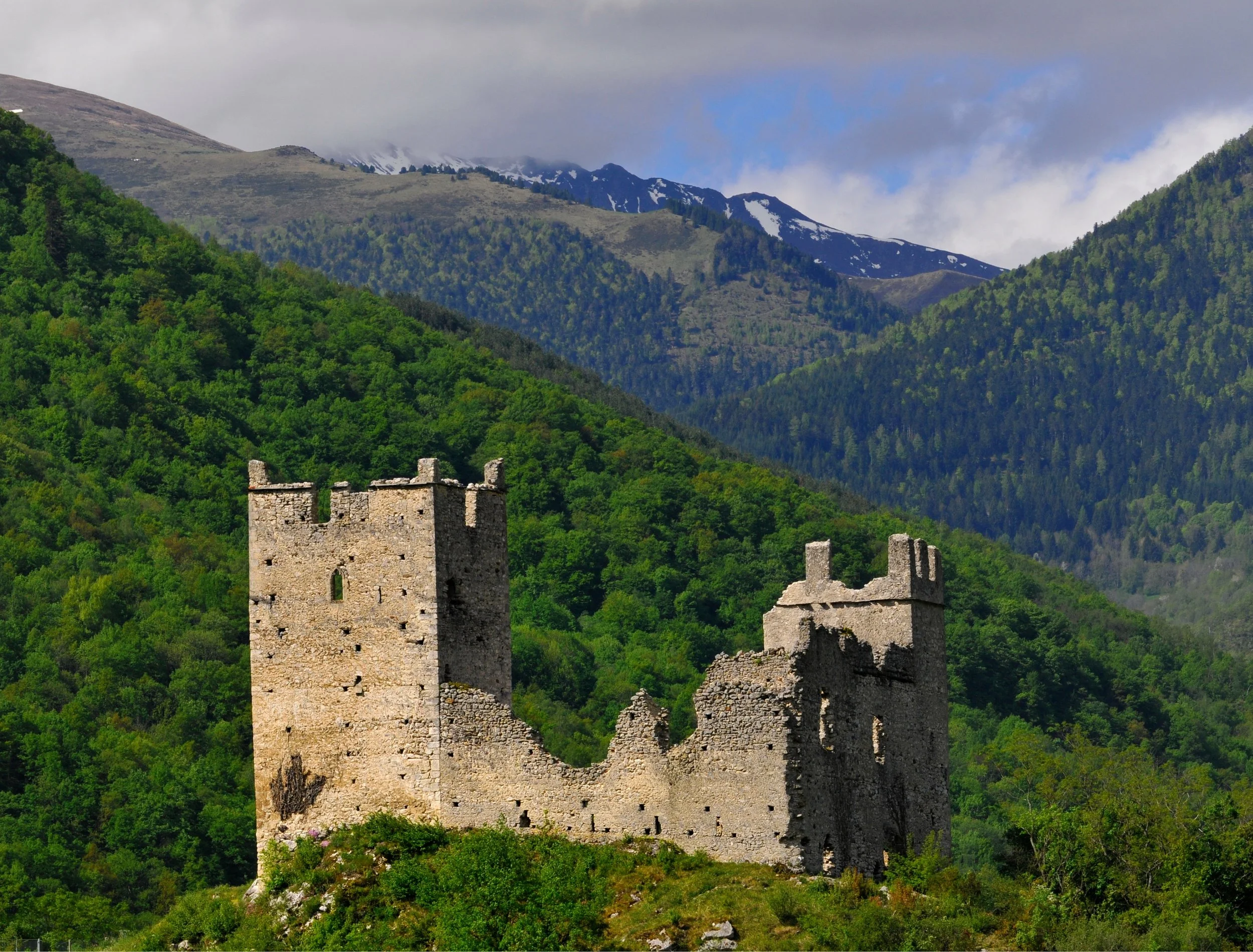 Ruins of an ancient stone castle with two towers, surrounded by green hills and forested mountains in the background under cloudy sky.