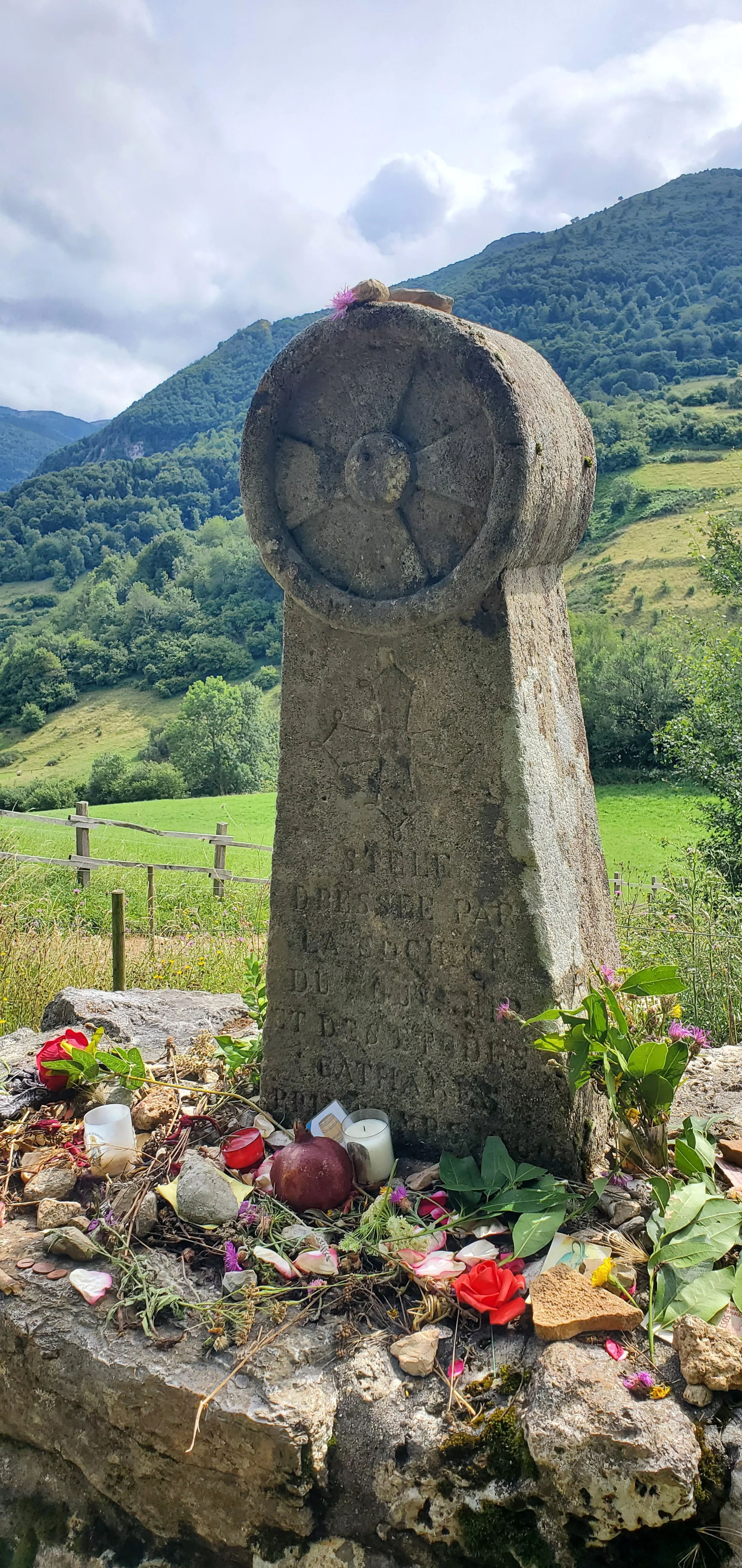 Stone memorial at Montsegur with flowers, candles, and offerings at the base, surrounded by green fields and hills under a partly cloudy sky.