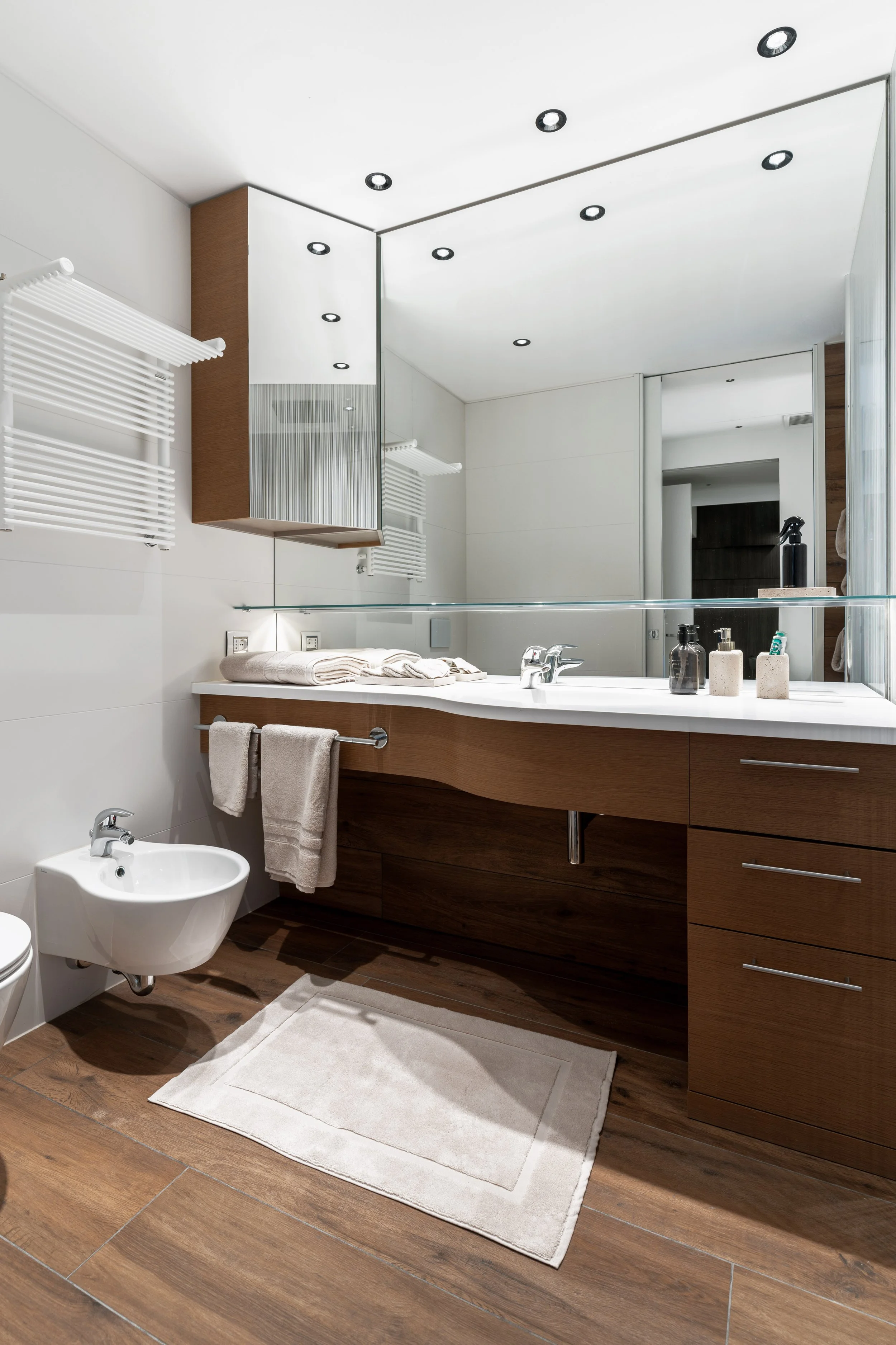 Bagno moderno con lavabo in legno, specchio grande, bidet, tendaggi bianchi, pavimento in legno, tappeto beige, prodotti per l'igiene e un rubinetto