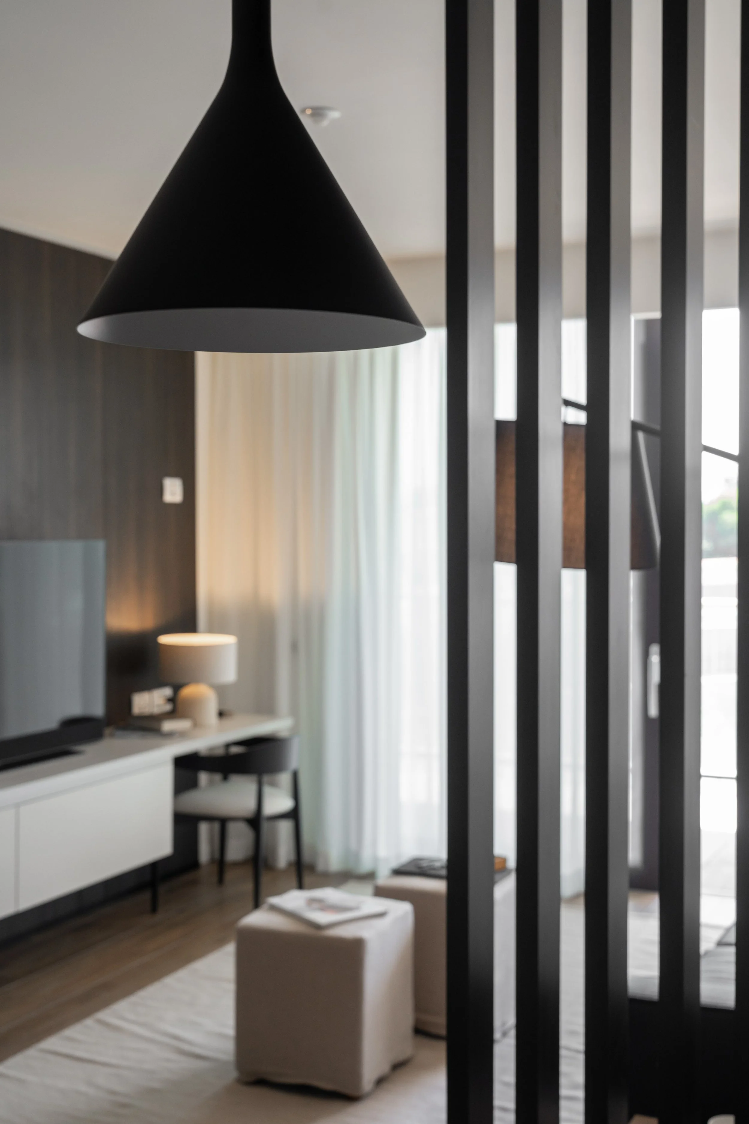 Interno di una stanza moderna con lampada nera a forma di cono, divanetto beige, televisore, tavolino, sedia nera, lampada da tavolo, tendaggi chiari, pareti scure e struttura di legno verticale.