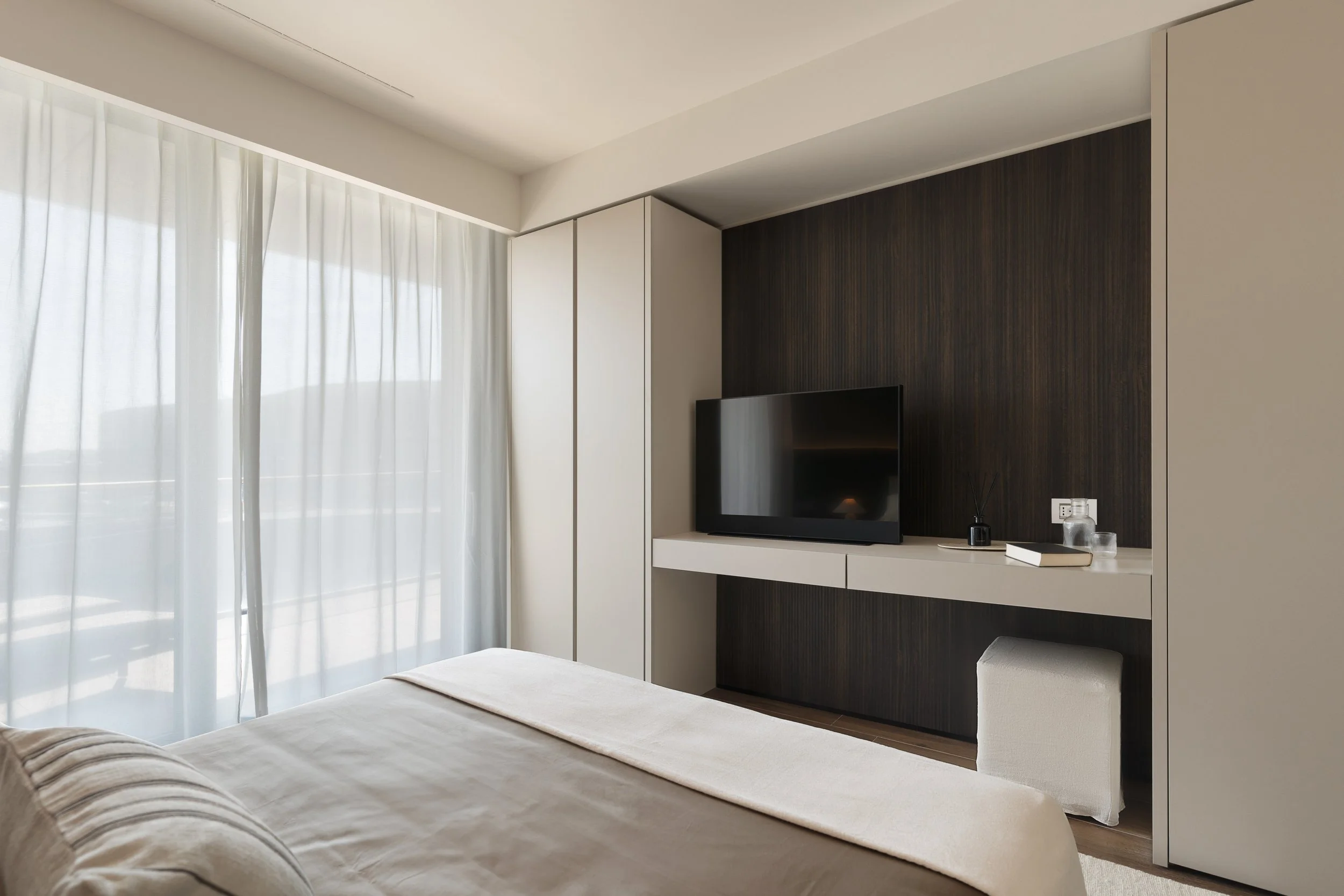Camera moderna con pareti bianche e scure, grande finestra con tende bianche, letto con lenzuola chiare, televisore su un mobile bianco, oggetti decorativi e prese di corrente vicino al televisore.